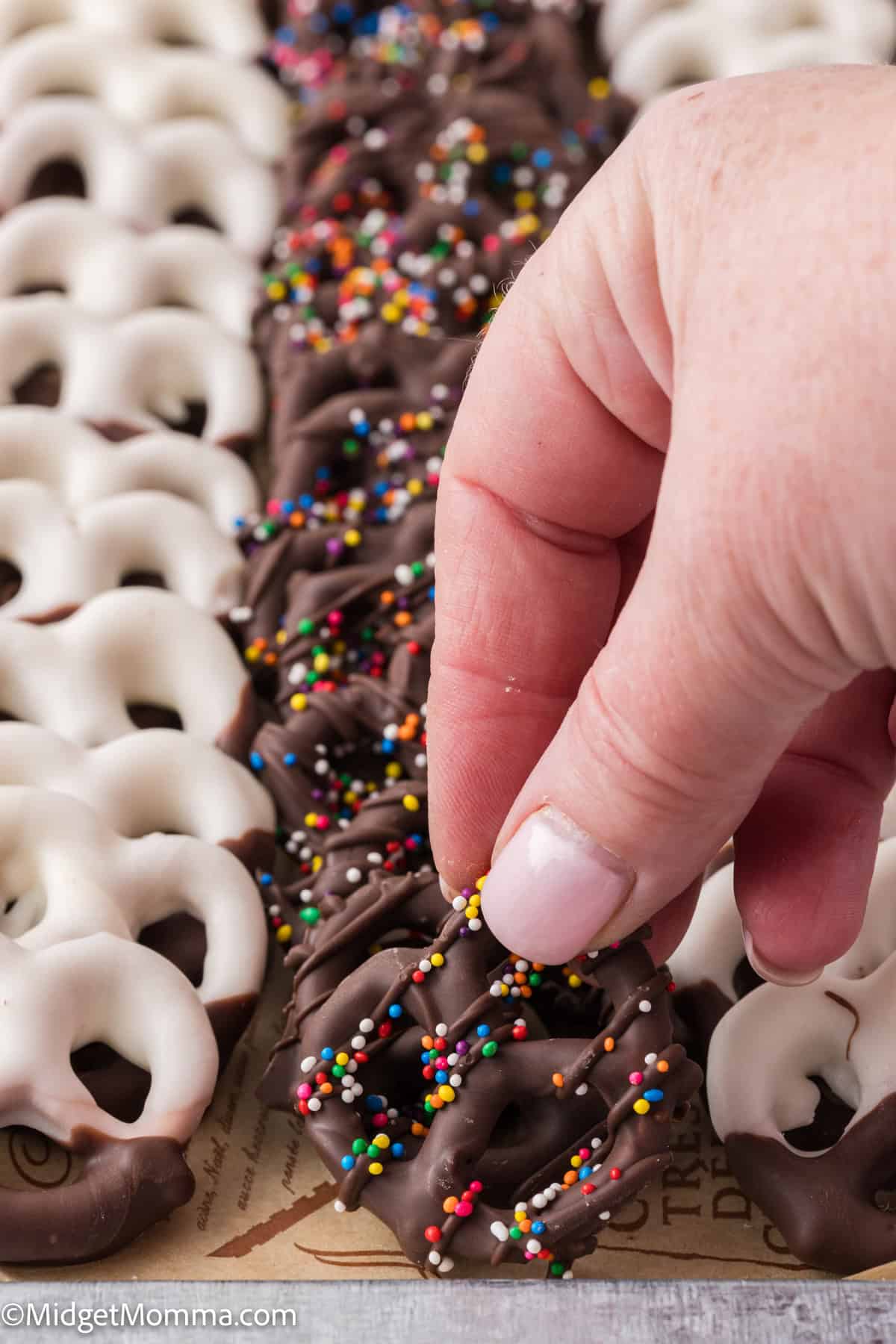 Hand holds a chocolate-covered pretzel with colorful sprinkles, next to white chocolate-covered pretzels on a tray.