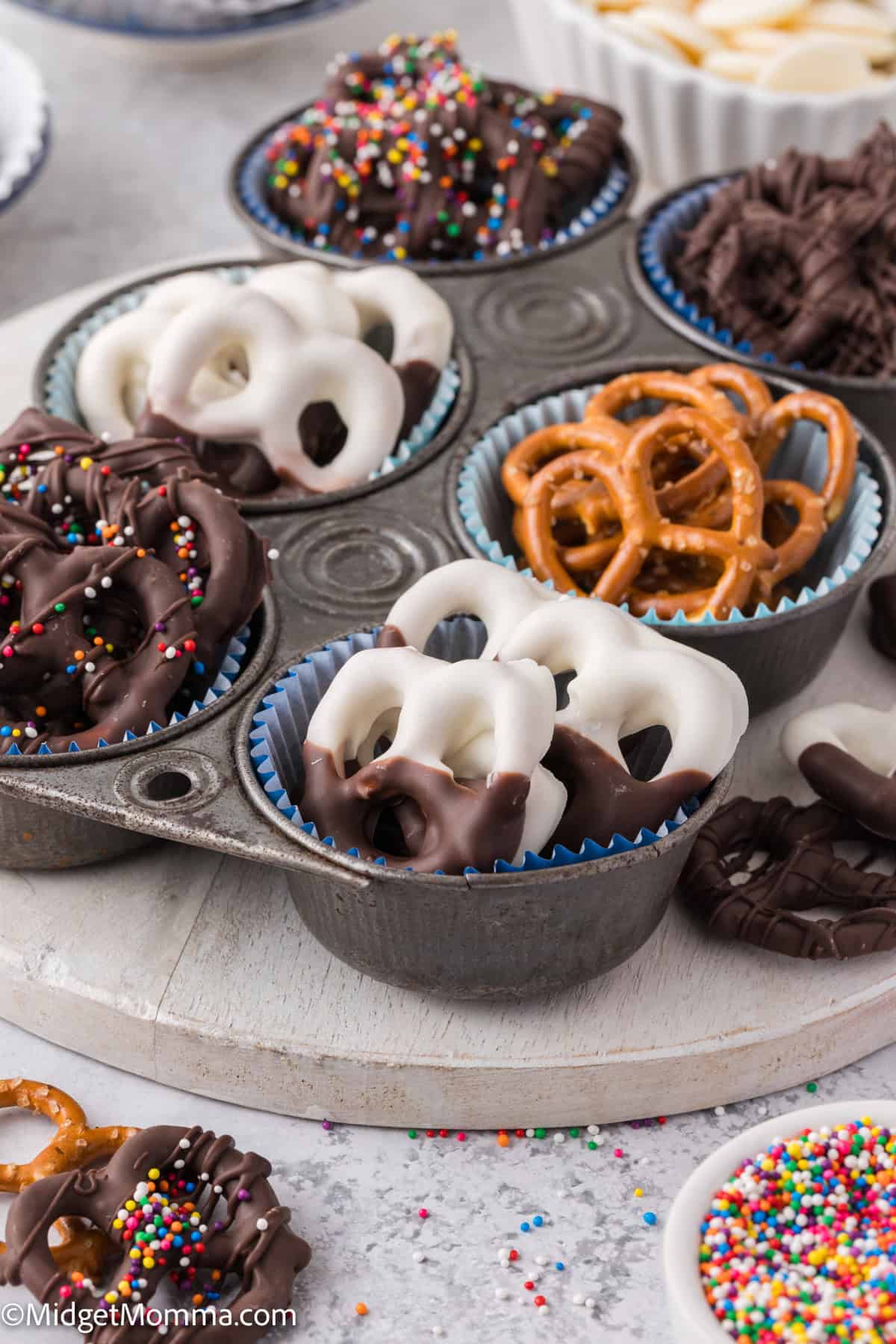 Assorted chocolate-covered pretzels with sprinkles displayed in muffin tin cups on a round tray.