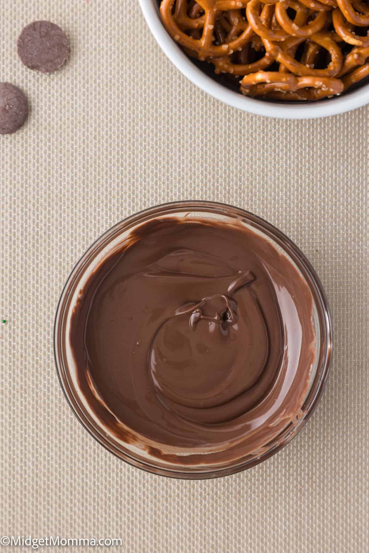 A bowl of melted chocolate next to a bowl of pretzels on a beige surface.