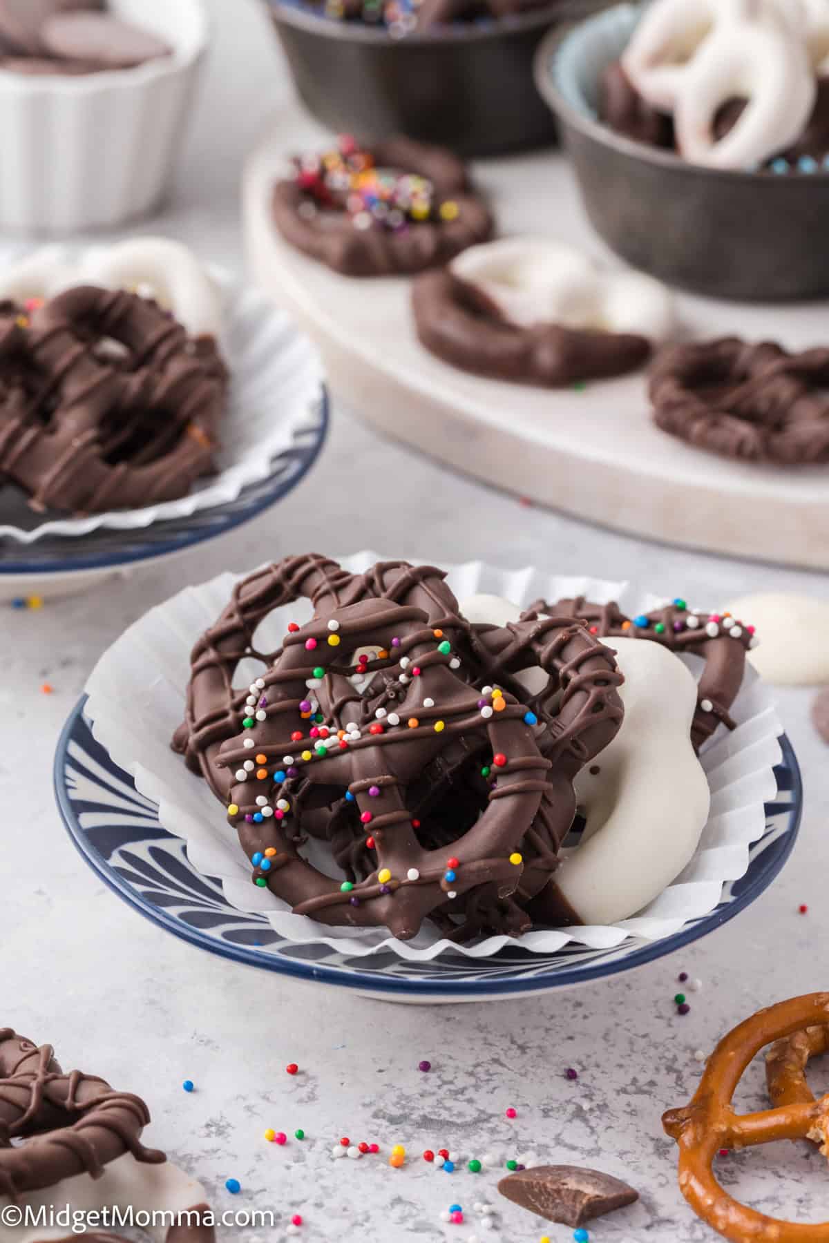 A plate of chocolate and yogurt-covered pretzels with colorful sprinkles, surrounded by more pretzels in bowls on a gray surface.