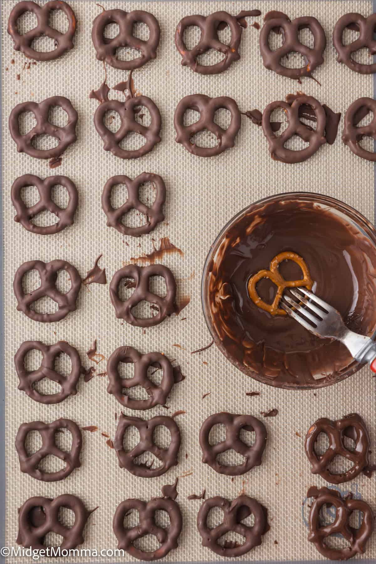 Pretzels dipped in chocolate arranged on a tray, with a bowl of melted chocolate and a fork holding another pretzel nearby.