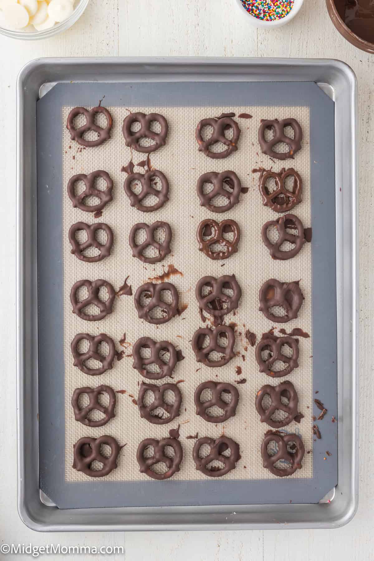 A baking tray with chocolate-covered pretzels arranged in rows on parchment paper, with a bowl of sprinkles partially visible in the corner.