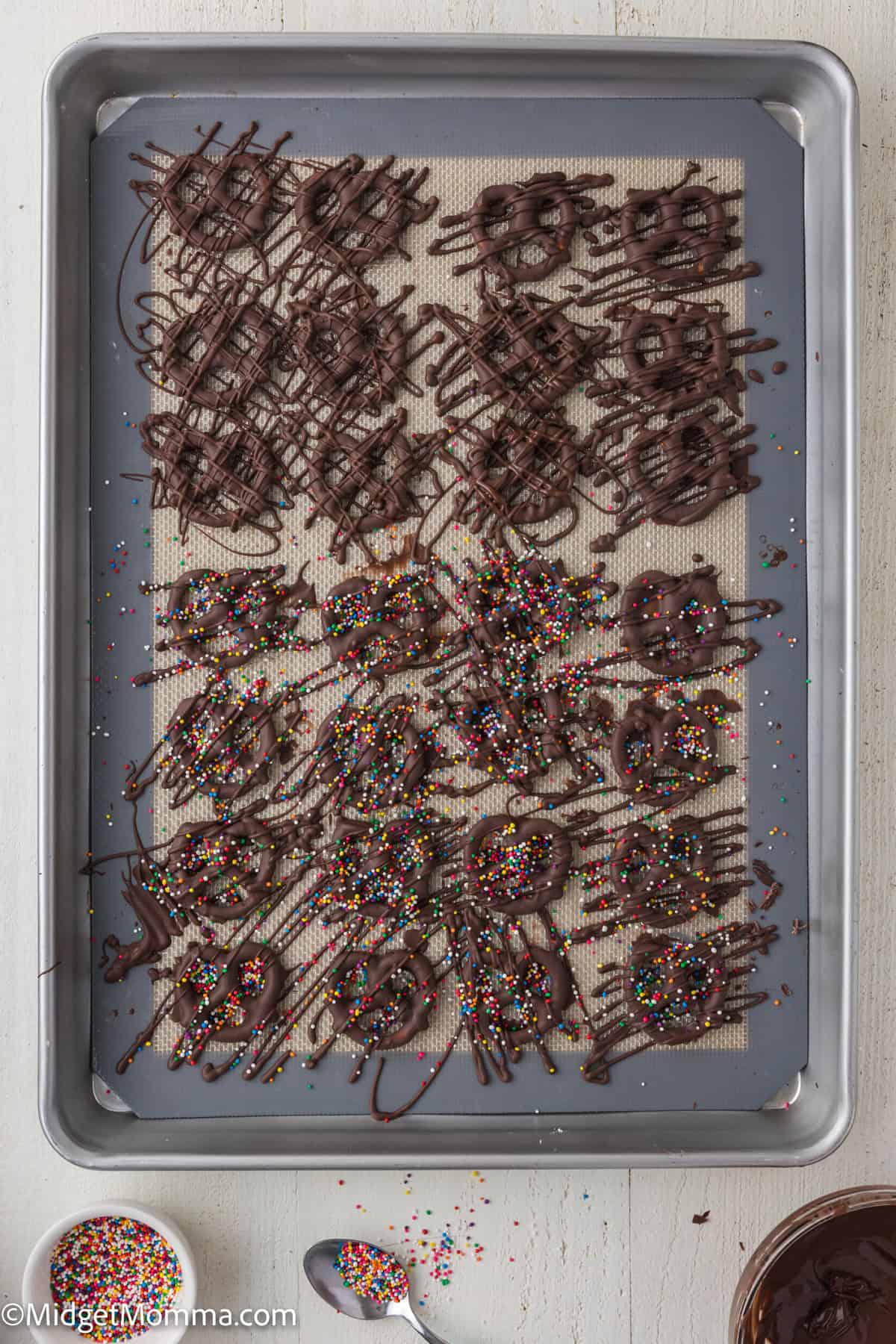 A baking sheet with chocolate-dipped pretzels drizzled with more chocolate and topped with colorful sprinkles.