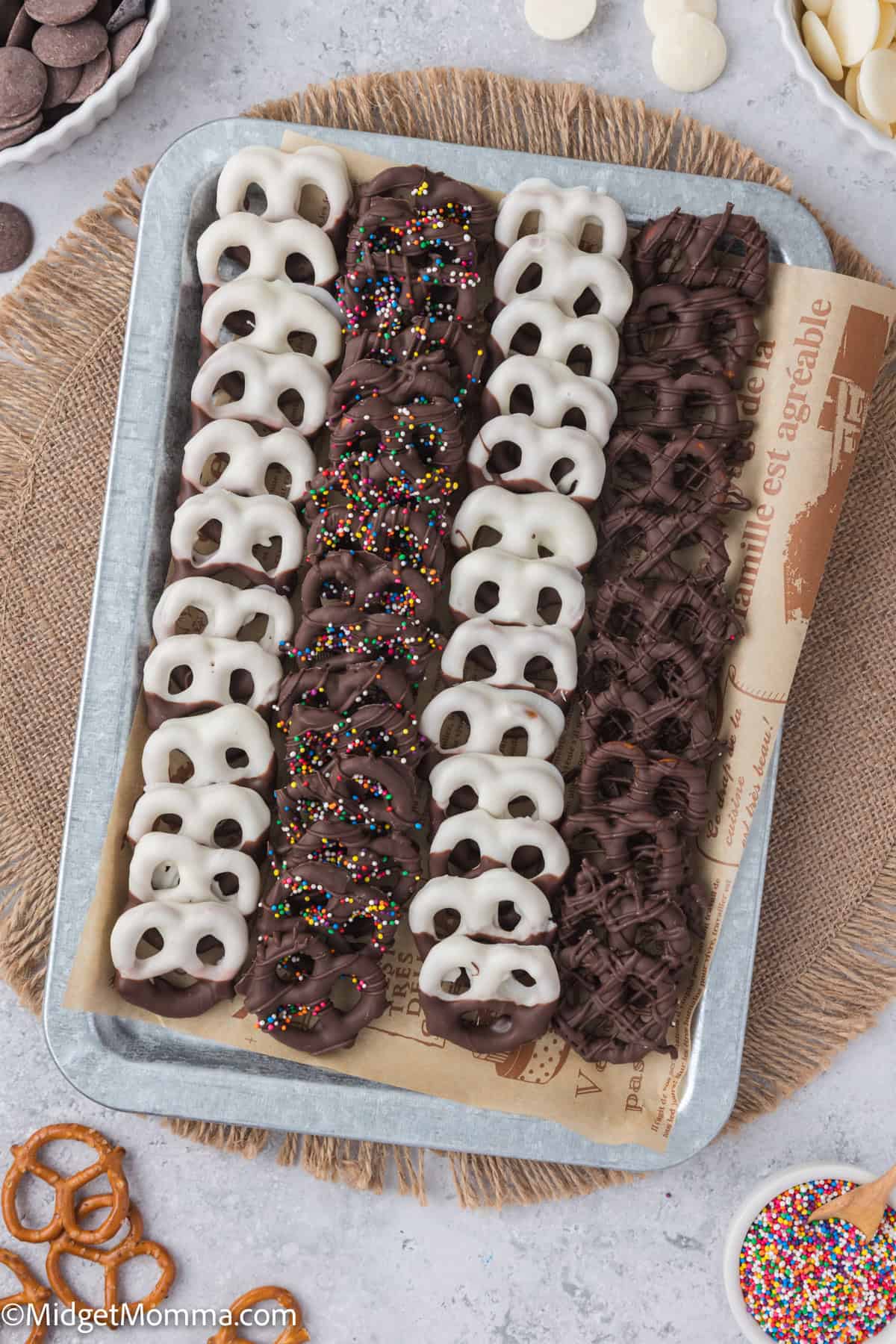 Tray with rows of white and milk chocolate-covered pretzels, some with sprinkles, on a burlap cloth.