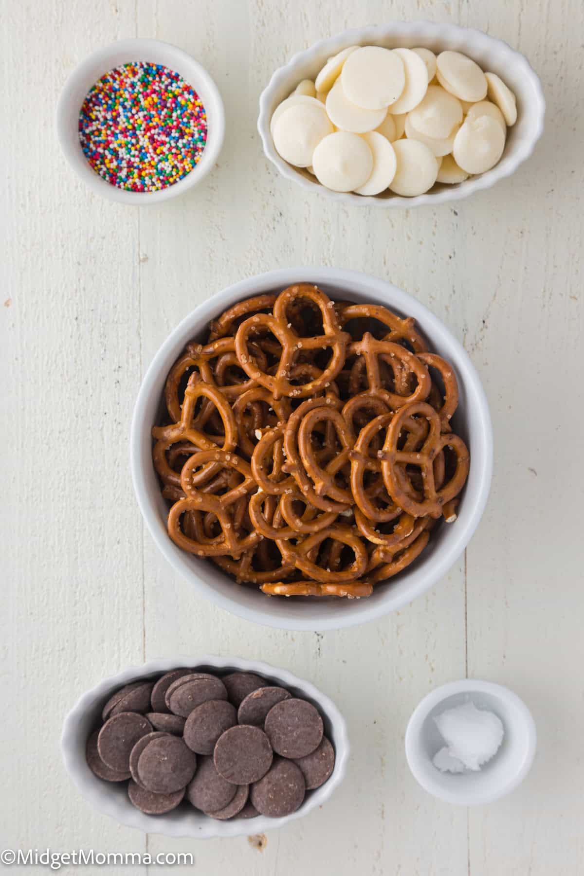 Bowls containing pretzels, white chocolate chips, dark chocolate chips, sprinkles, and a small bowl of salt on a light surface.