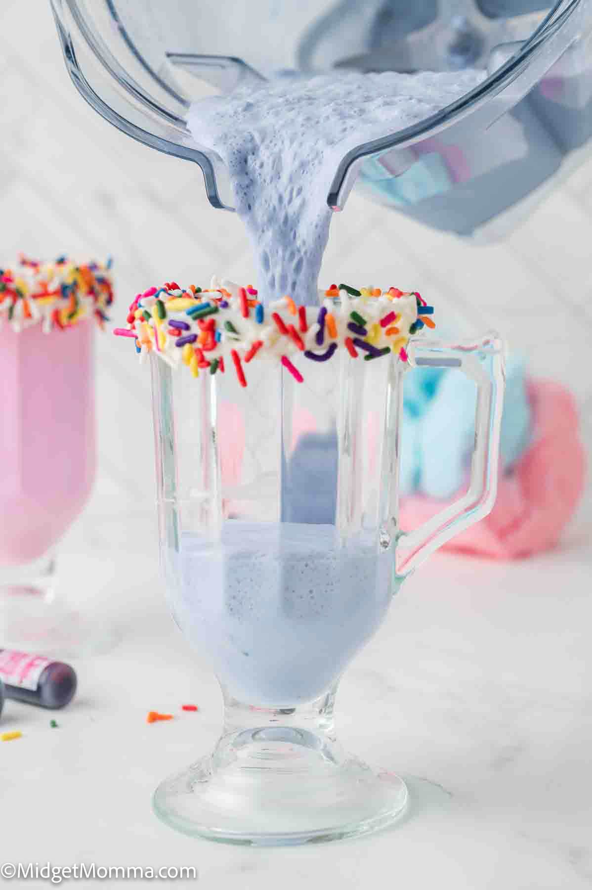 A blue milkshake is being poured from a blender into a glass with a rim coated in frosting and colorful sprinkles.