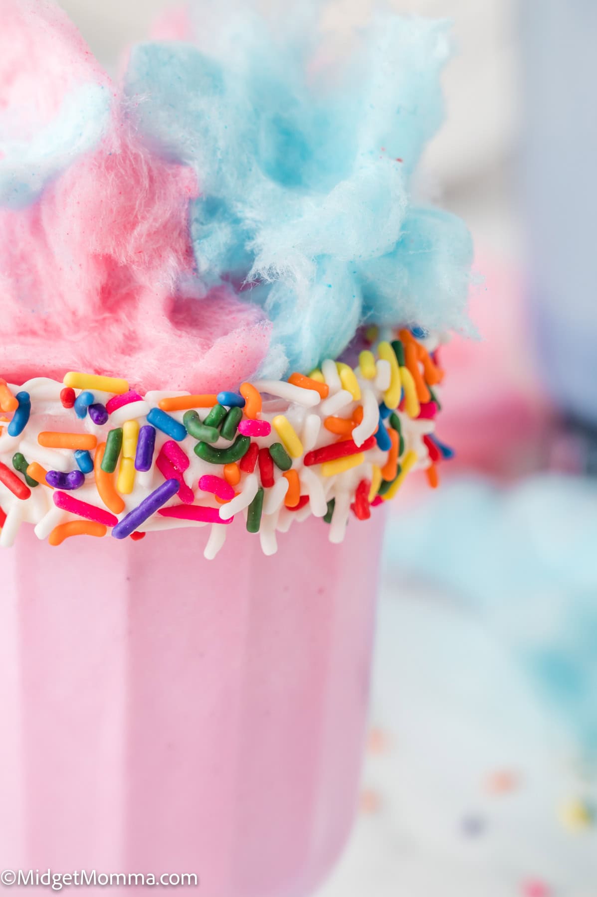 A pink milkshake in a glass rimmed with colorful sprinkles, topped with pink and blue cotton candy.