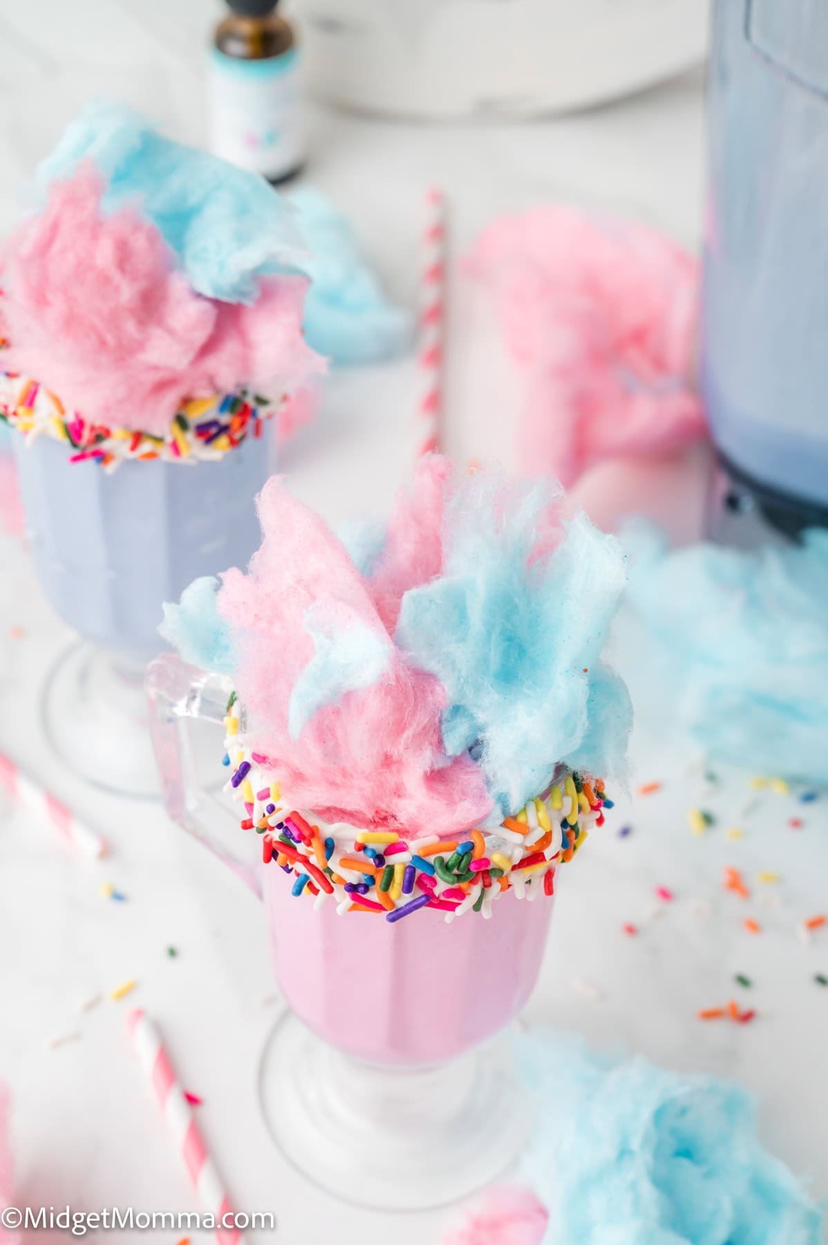 A pastel pink milkshake in a glass with sprinkles on the rim, topped with pink and blue cotton candy. Another similar glass is partially visible in the background.