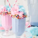 Two pastel milkshakes in clear glasses, topped with cotton candy and sprinkles, each with a striped straw; a pitcher of milkshake and cotton candy pieces are in the background.