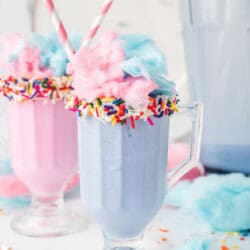 Two pastel milkshakes in clear glasses, topped with cotton candy and sprinkles, each with a striped straw; a pitcher of milkshake and cotton candy pieces are in the background.