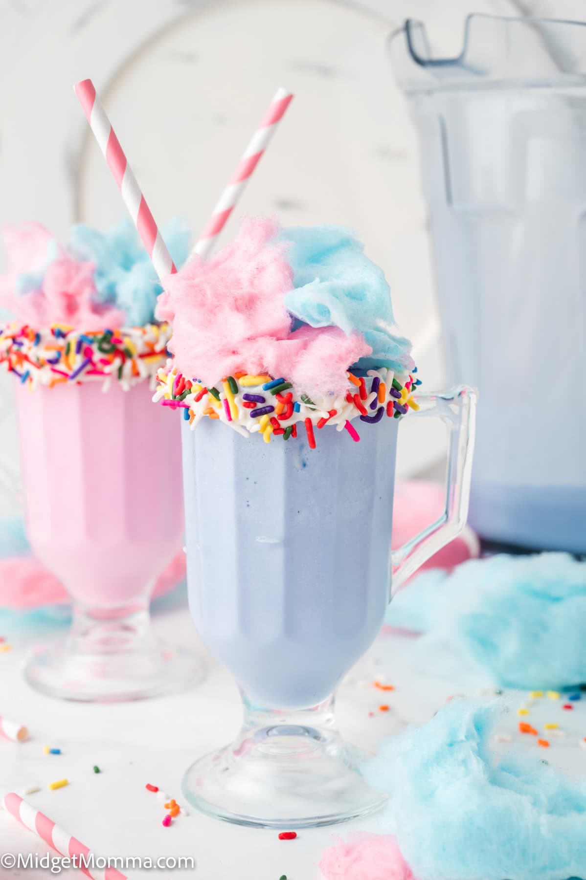 Two pastel milkshakes in clear glasses, topped with cotton candy and sprinkles, each with a striped straw; a pitcher of milkshake and cotton candy pieces are in the background.