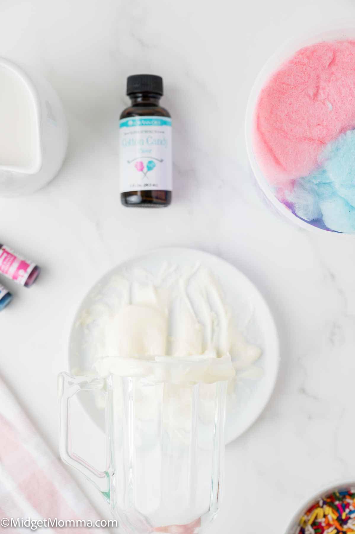 A blender with vanilla ice cream, a bottle of cotton candy flavoring, a bowl of pink and blue cotton candy, and colorful sprinkles on a white surface.