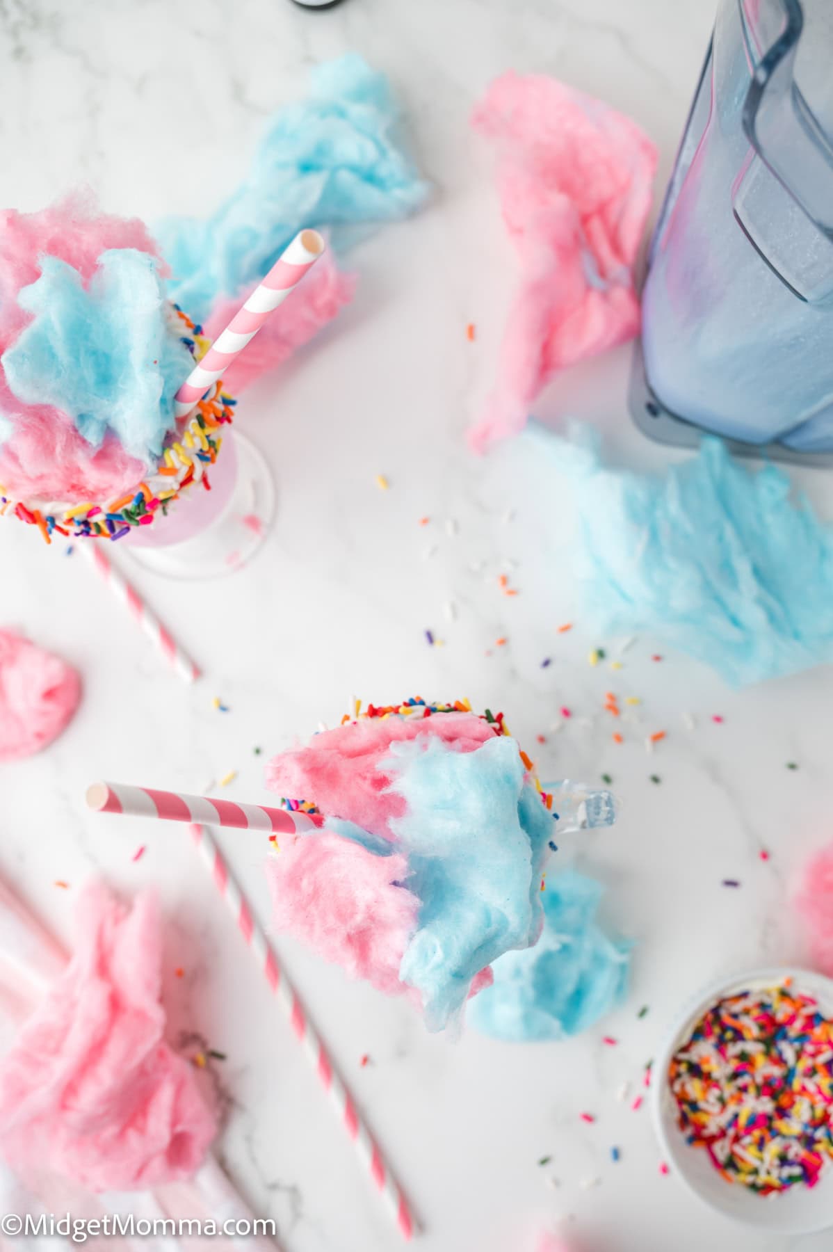 Two glasses topped with pink and blue cotton candy and rainbow sprinkles, surrounded by scattered cotton candy, sprinkles, and a blender on a white surface.