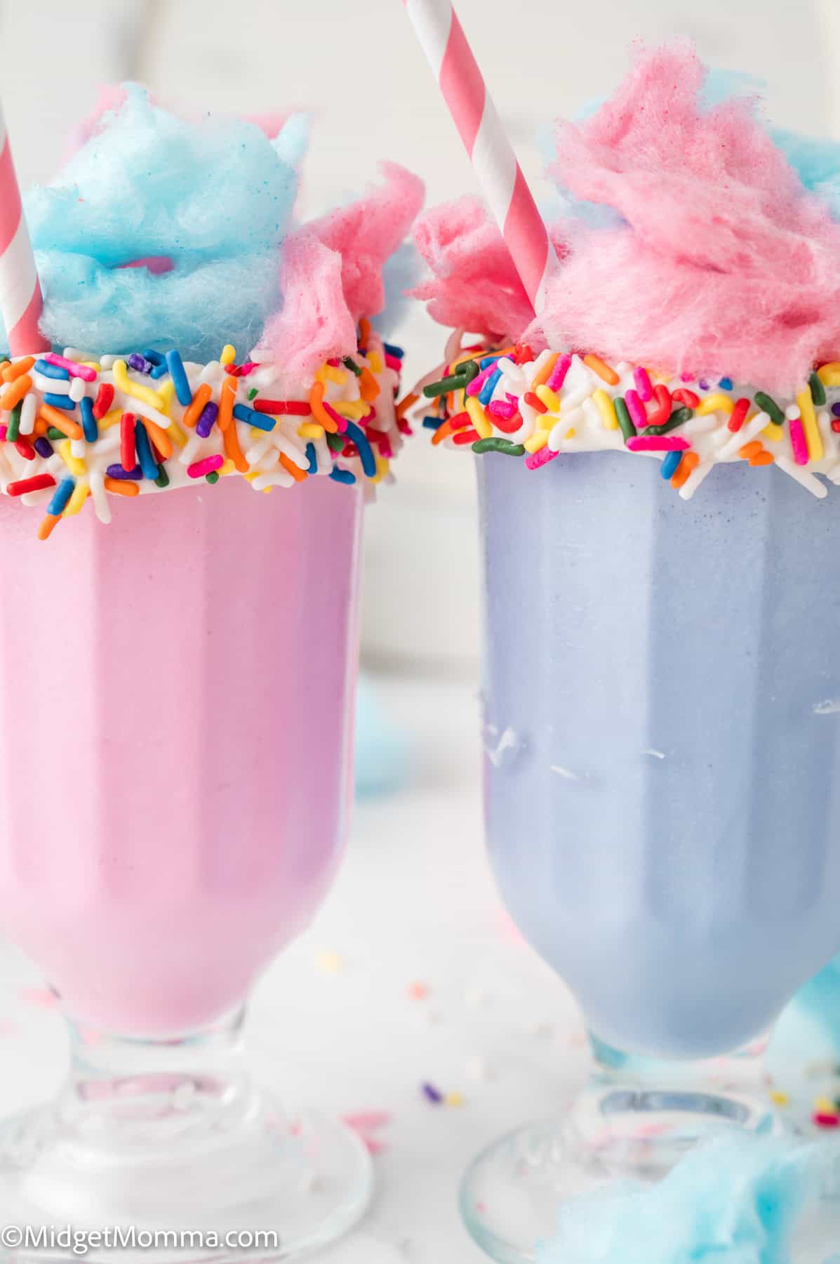Two milkshakes, one pink and one light purple, topped with blue and pink cotton candy and rainbow sprinkles, served in clear glasses with striped straws.