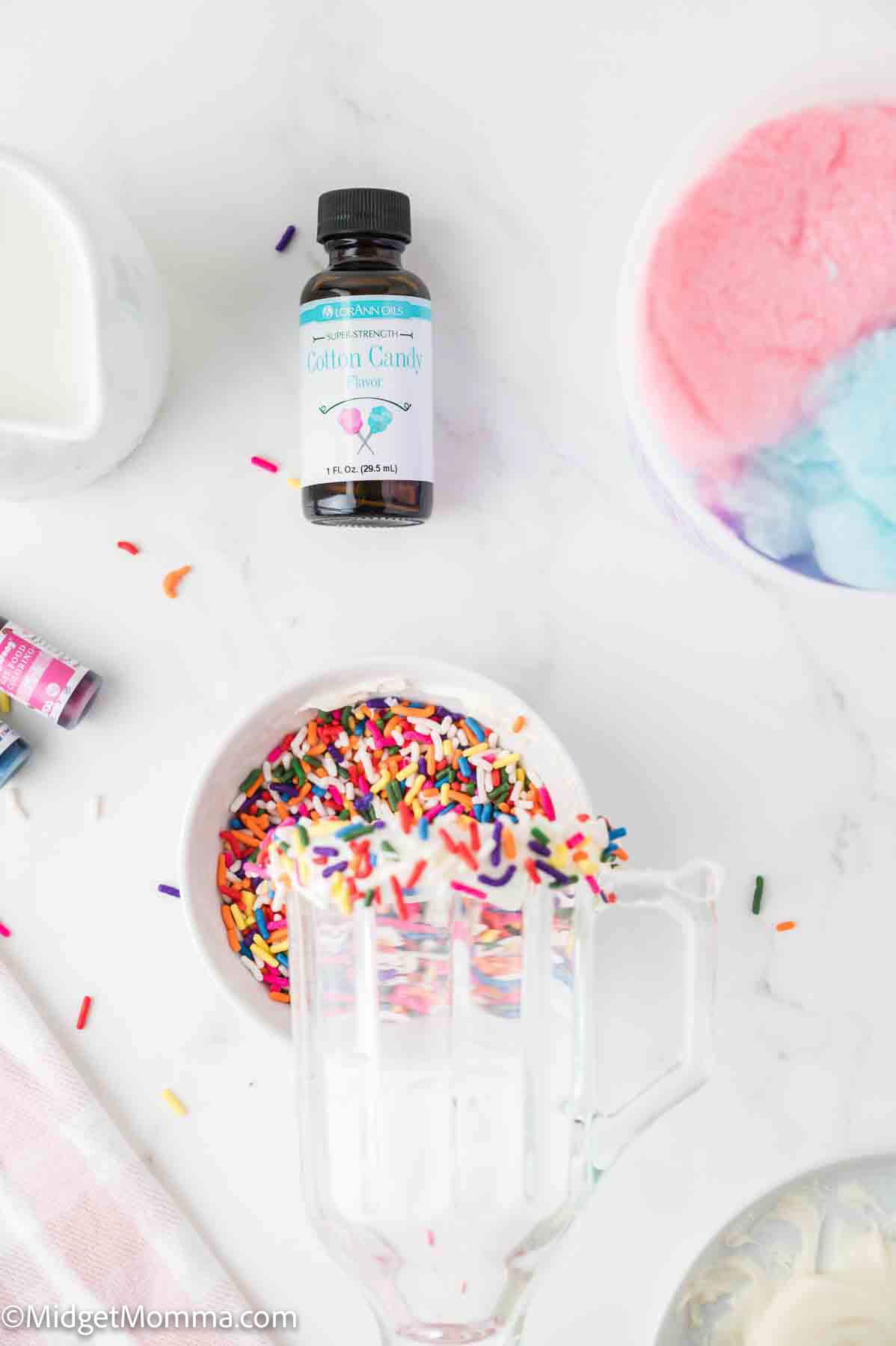 A bottle of cotton candy flavoring, a bowl of colorful sprinkles, and a bowl of cotton candy on a white surface with a glass measuring cup.