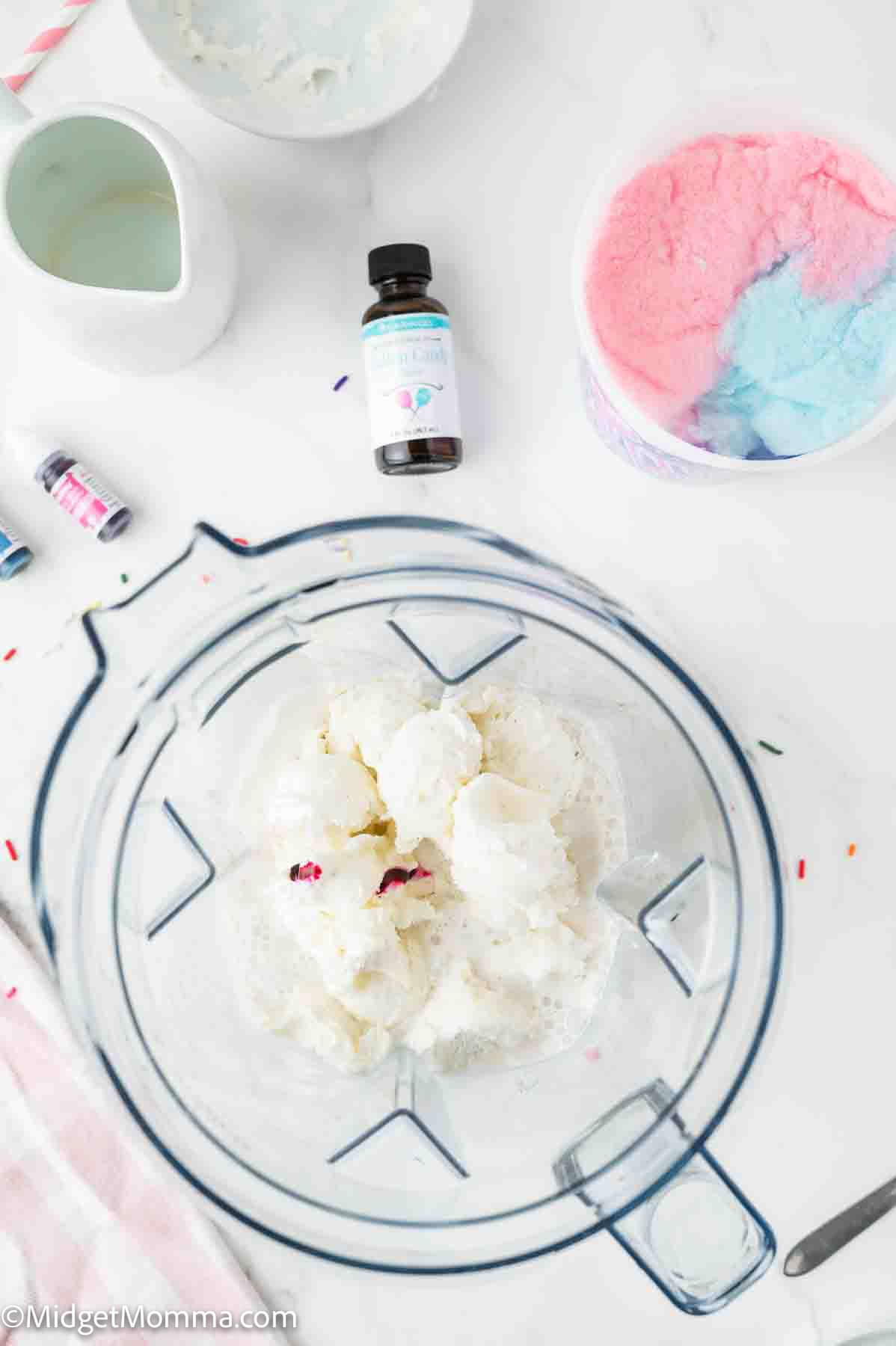 A blender with scoops of vanilla ice cream, a bowl of pink and blue cotton candy, food coloring bottles, and a bottle of flavoring on a white countertop.