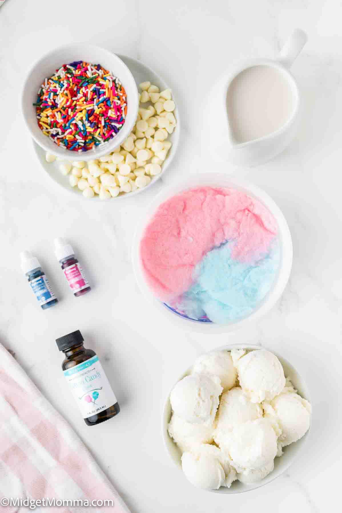 Top-down view of bowls containing rainbow sprinkles, white chocolate chips, milk, cotton candy, scoops of ice cream, food coloring, and a bottle of extract on a white surface.