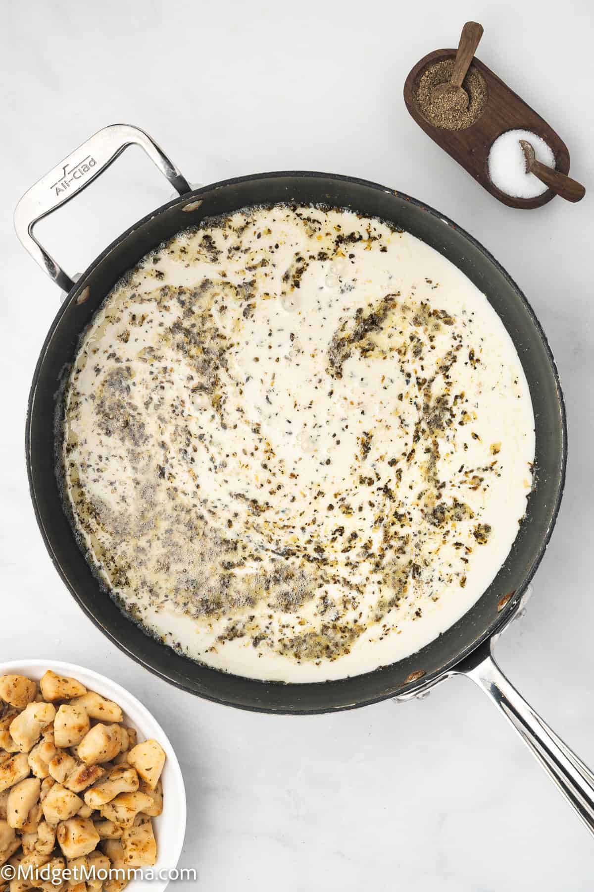 A skillet filled with creamy sauce and herbs sits on a white surface, with a bowl of cooked chicken pieces and a dish of salt and pepper nearby.