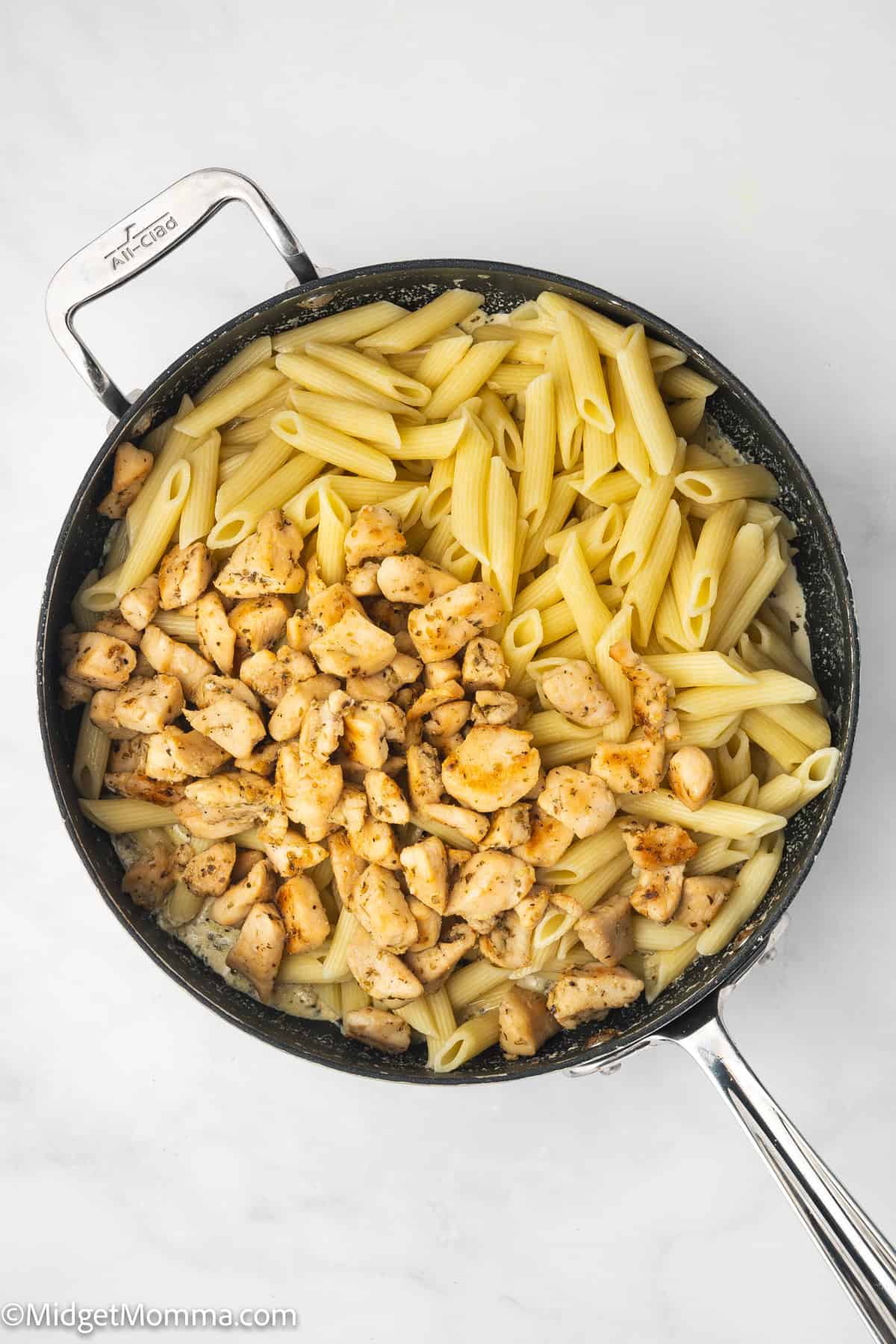 A skillet containing cooked penne pasta and seasoned diced chicken pieces, separated, on a white surface.