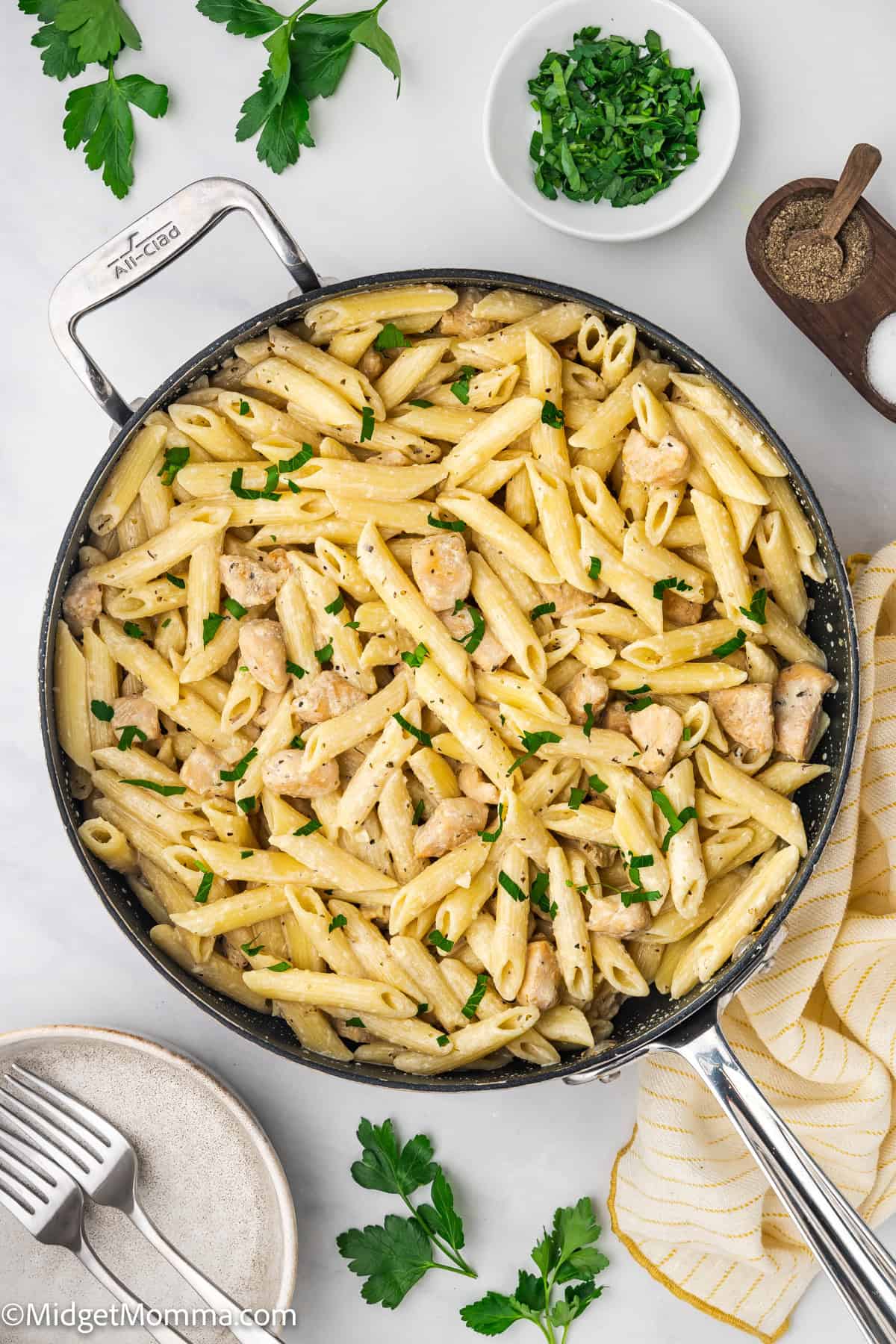 A skillet filled with penne pasta and chicken in a light sauce, garnished with chopped parsley, sits on a white surface with bowls of parsley and pepper nearby.