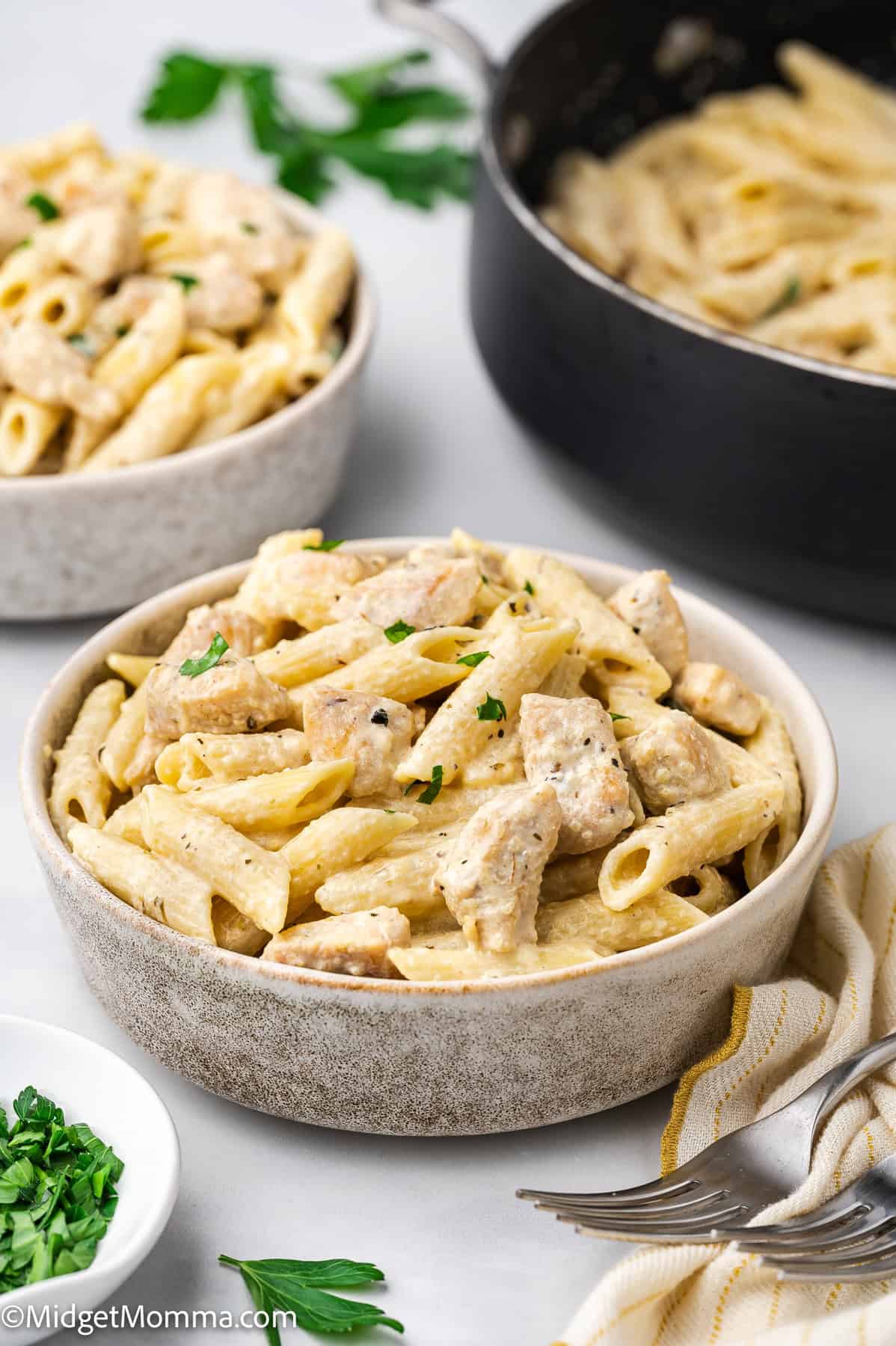 A bowl of creamy penne pasta with chunks of chicken, garnished with chopped parsley, sits on a table next to a pan and another bowl of the same dish.