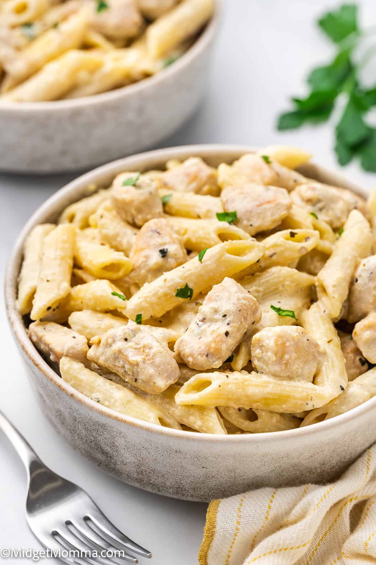 A bowl of penne pasta with creamy sauce and pieces of chicken, garnished with chopped herbs, sits on a white surface next to a fork and cloth napkin.