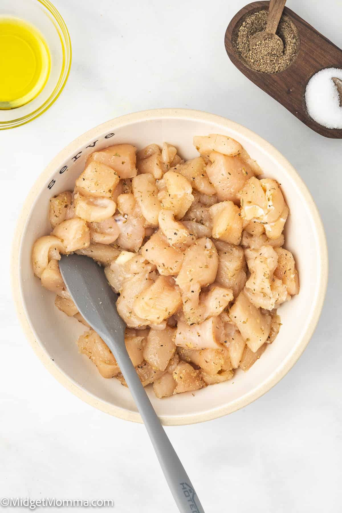 A bowl of raw, diced chicken seasoned with salt and pepper, with a spatula in the bowl. A small bowl of olive oil and a tray of salt and pepper are nearby.