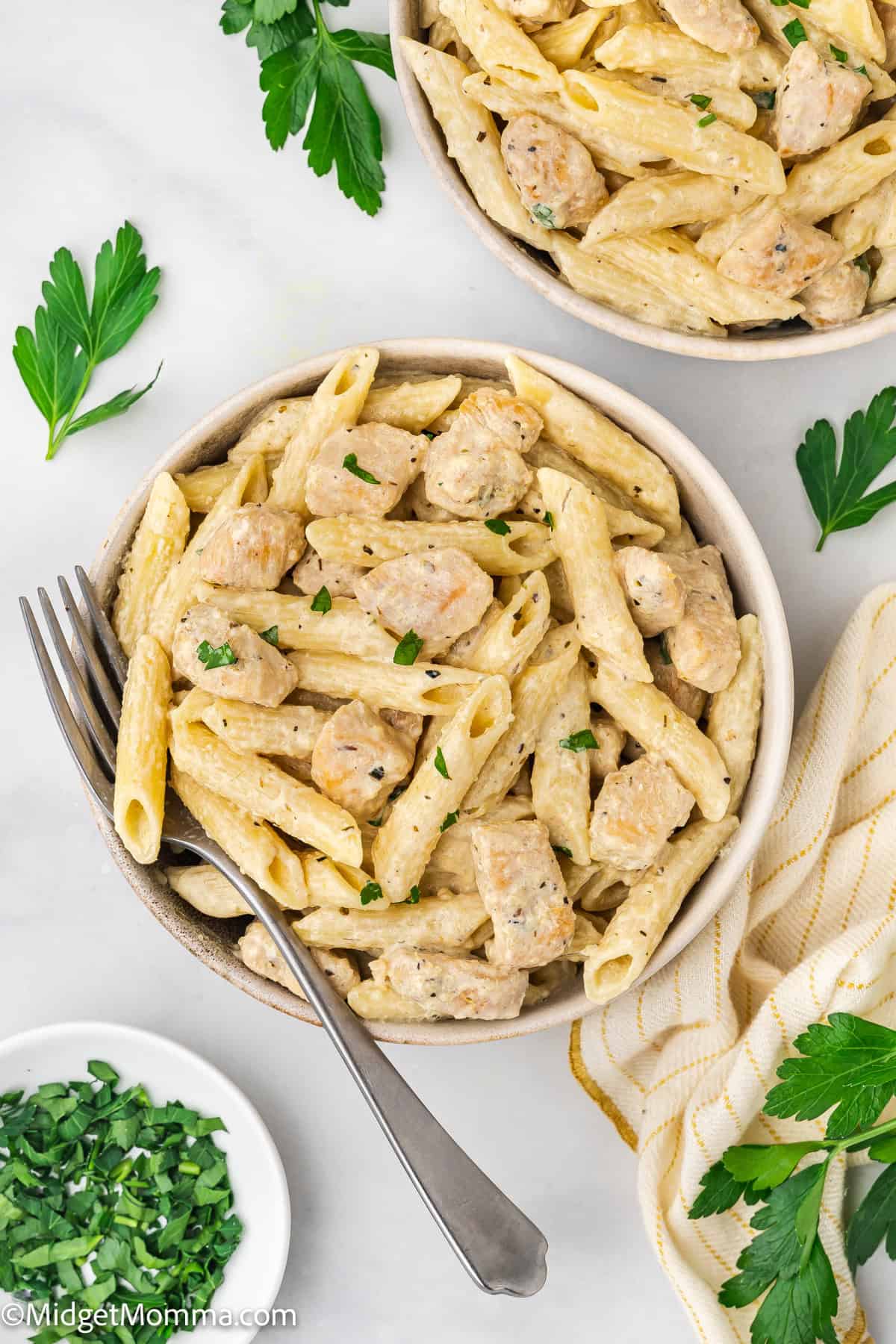 A bowl of creamy penne pasta with chicken pieces, garnished with chopped parsley, sits on a white surface next to a fork and a small dish of herbs.