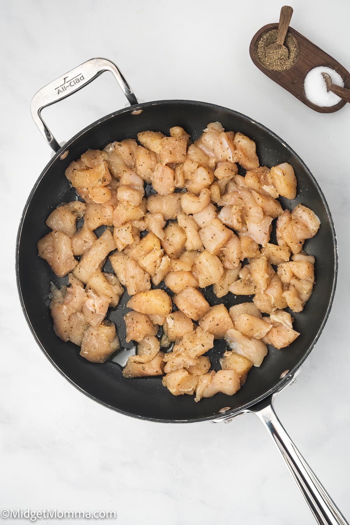 Diced, seasoned chicken pieces cooking in a black frying pan on a white surface, with salt and pepper in a small wooden dish nearby.