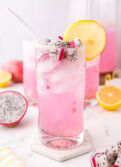 A glass of pink dragon fruit lemonade with ice, topped with dragon fruit pieces and a lemon slice. Two similar glasses and fruit halves are in the background.