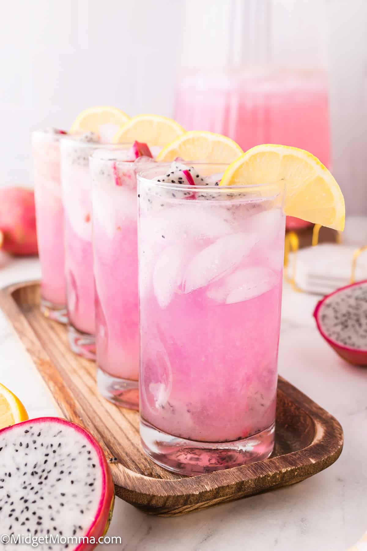 Four glasses of pink dragon fruit lemonade with ice and lemon slices on a wooden tray, surrounded by dragon fruit halves.