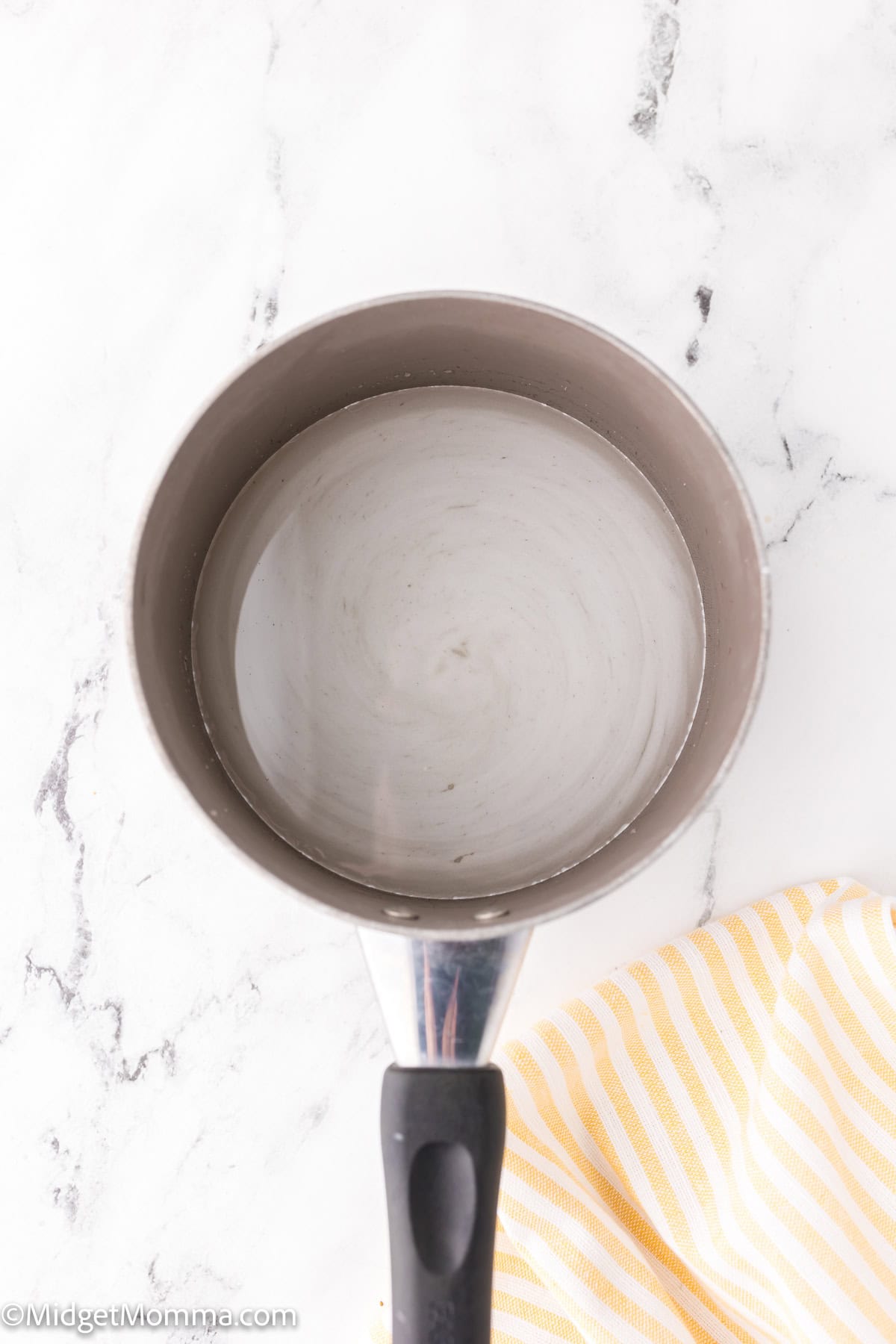 A pot with a black handle containing a clear liquid, possibly water, is placed on a marble surface next to a yellow and white striped cloth.