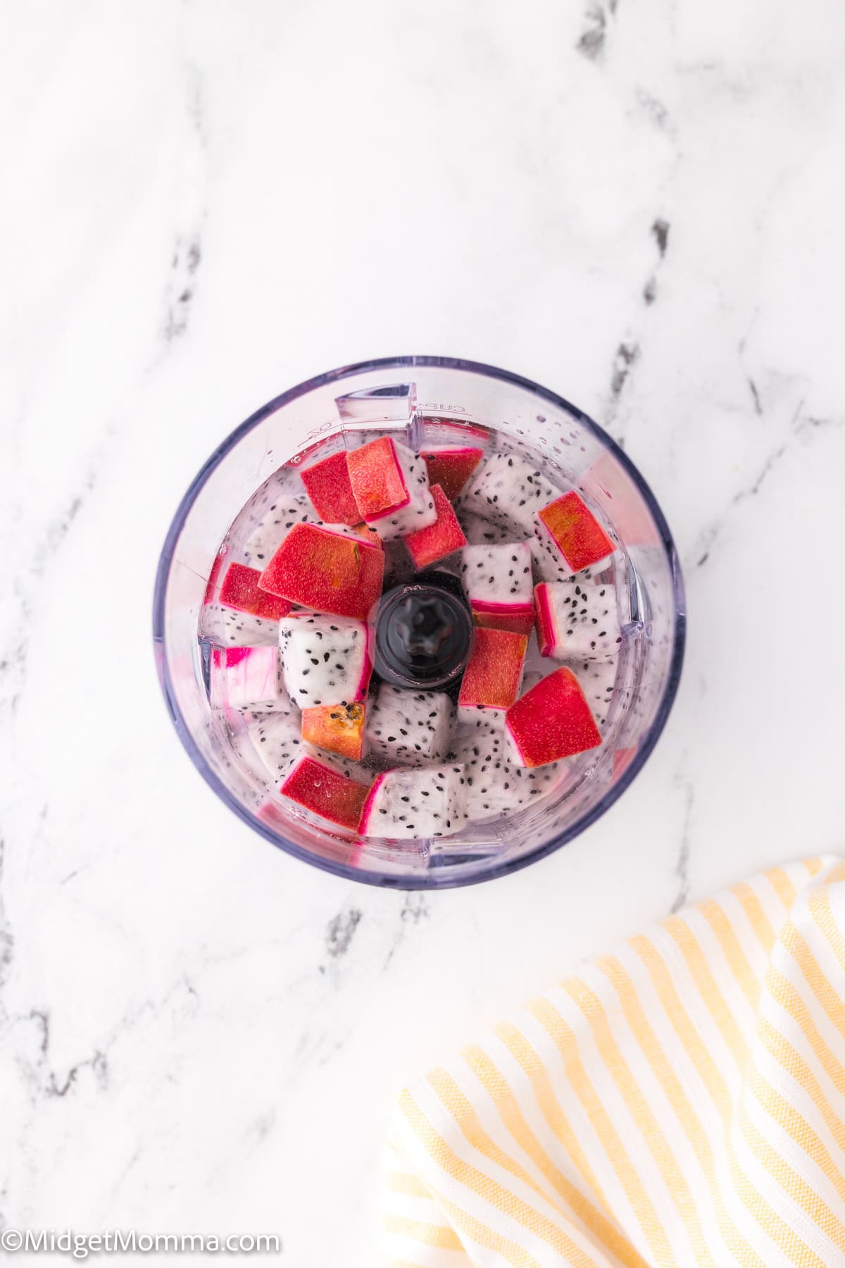 Top view of a food processor containing chopped red and white dragon fruit cubes on a marbled surface. A striped cloth is partially visible in the corner.