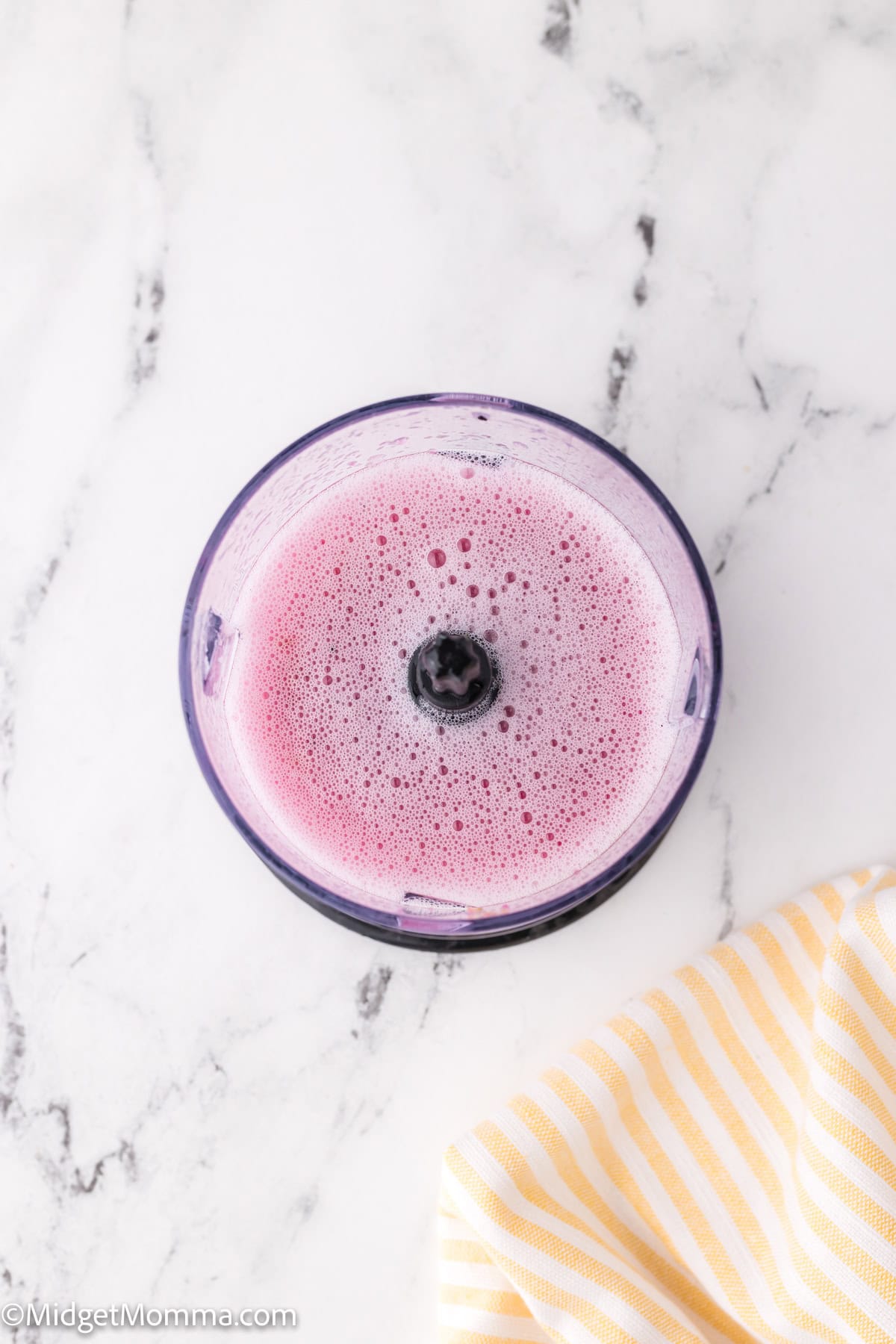 Top view of a blender cup with pink smoothie and a blueberry on top, on a marble surface next to a yellow striped cloth.