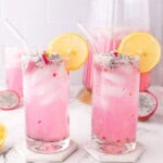 Two tall glasses of pink dragon fruit lemonade with ice, garnished with lemon slices and dragon fruit cubes. A pitcher of the drink and sliced fruit are in the background on a marble surface.