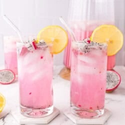 Two tall glasses of pink dragon fruit lemonade with ice, garnished with lemon slices and dragon fruit cubes. A pitcher of the drink and sliced fruit are in the background on a marble surface.