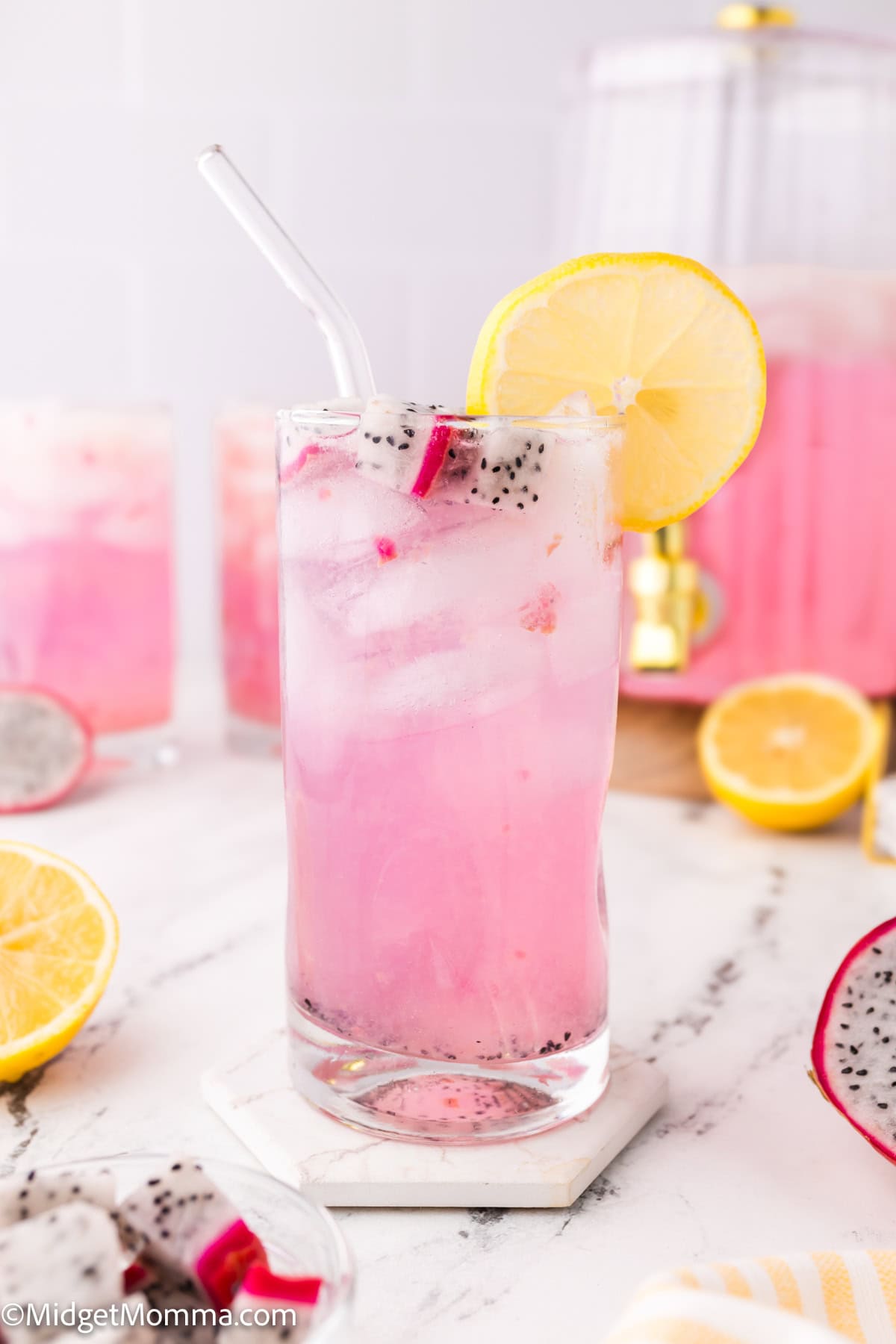 Glass of pink dragon fruit lemonade with ice, a lemon slice on the rim, and a straw. Additional lemon and dragon fruit slices nearby on a marble surface.