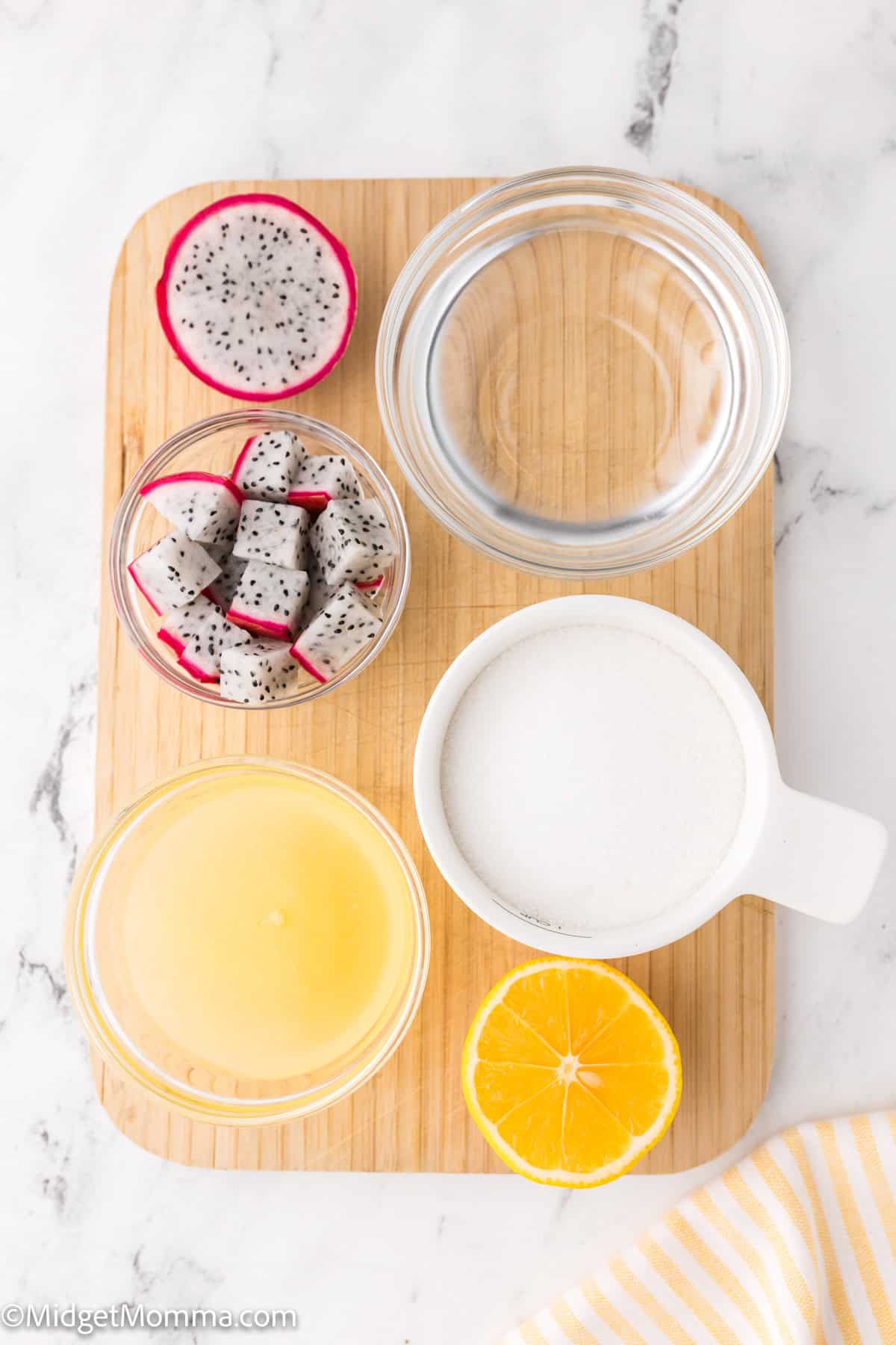 Wooden board with bowls of dragon fruit pieces, sugar, and water. There's also a glass of lemon juice and a half lemon on a marble surface.