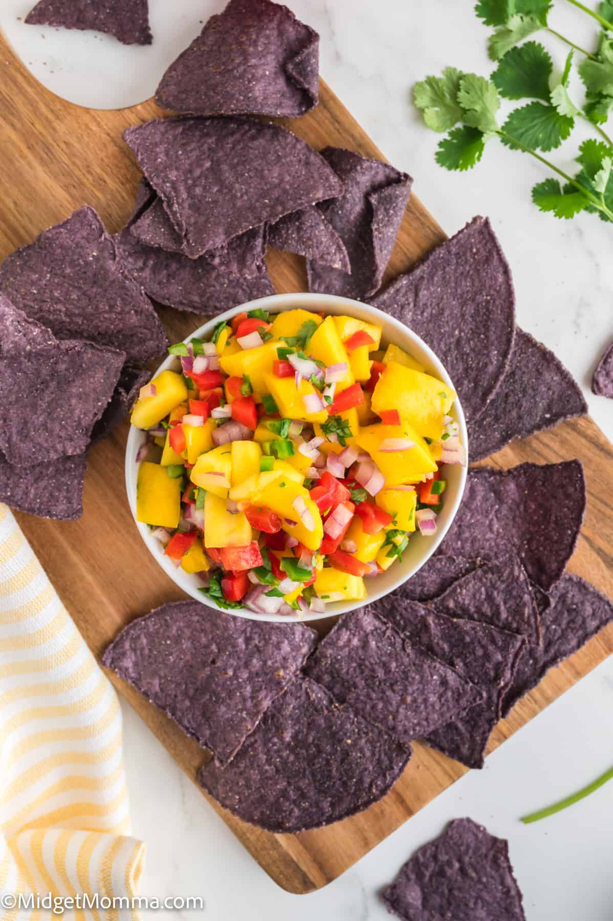 A bowl of mango salsa with diced red onion, tomato, and jalapeño is surrounded by blue corn tortilla chips on a wooden board.