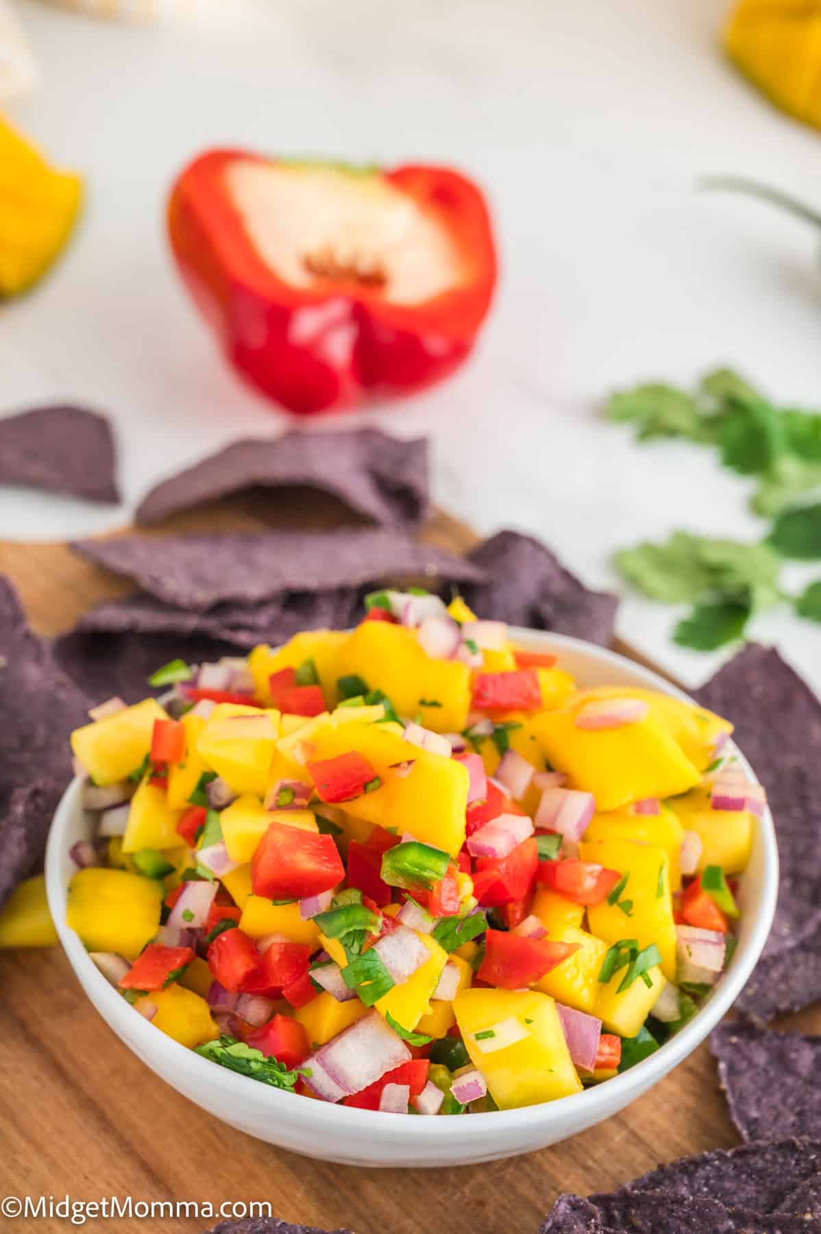 A bowl of mango salsa with diced mango, red onion, tomato, and cilantro is surrounded by blue corn tortilla chips. A halved red bell pepper is in the background.