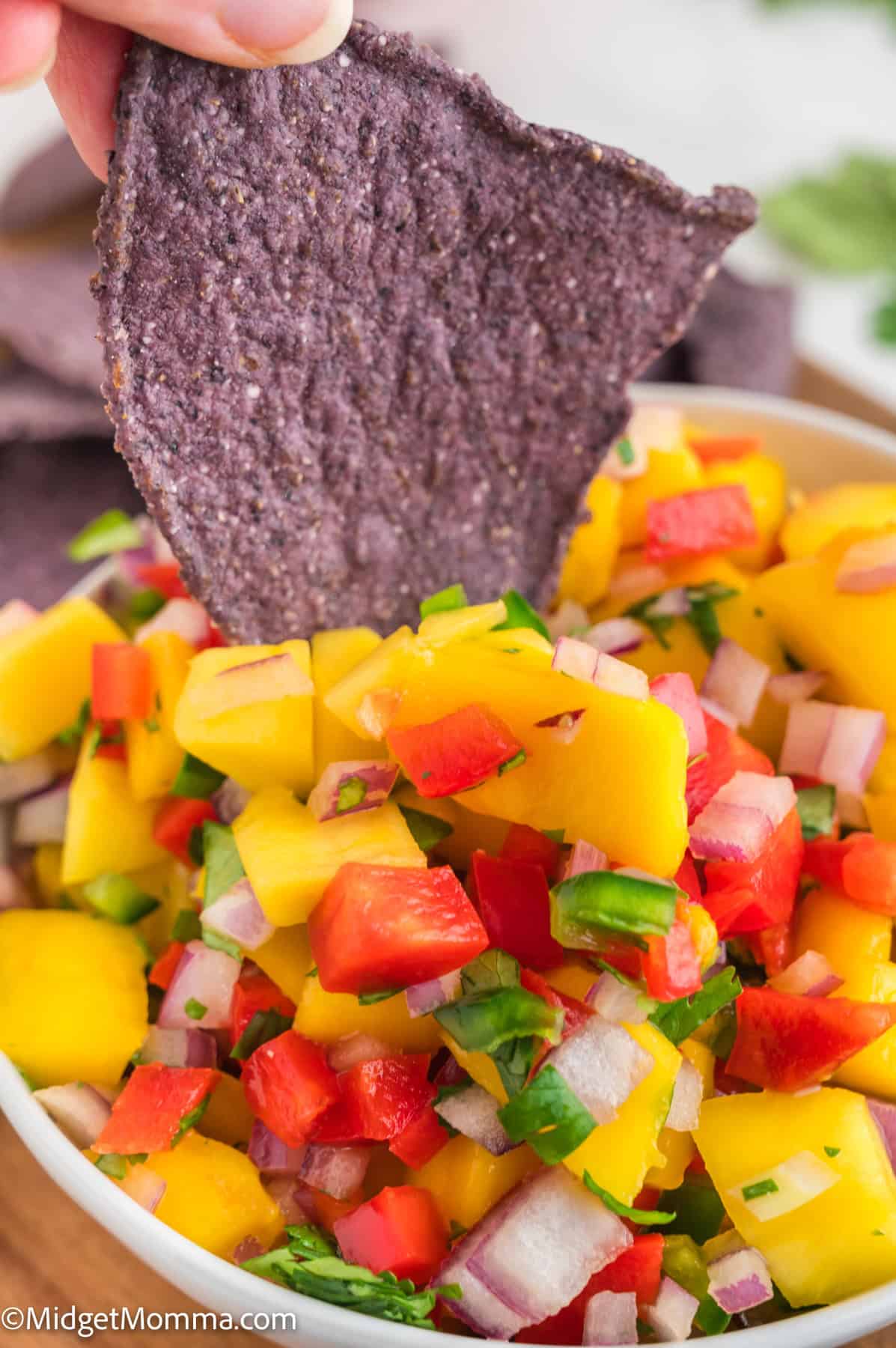 A hand dips a blue corn tortilla chip into a bowl of mango salsa with chopped red pepper, onion, and cilantro.