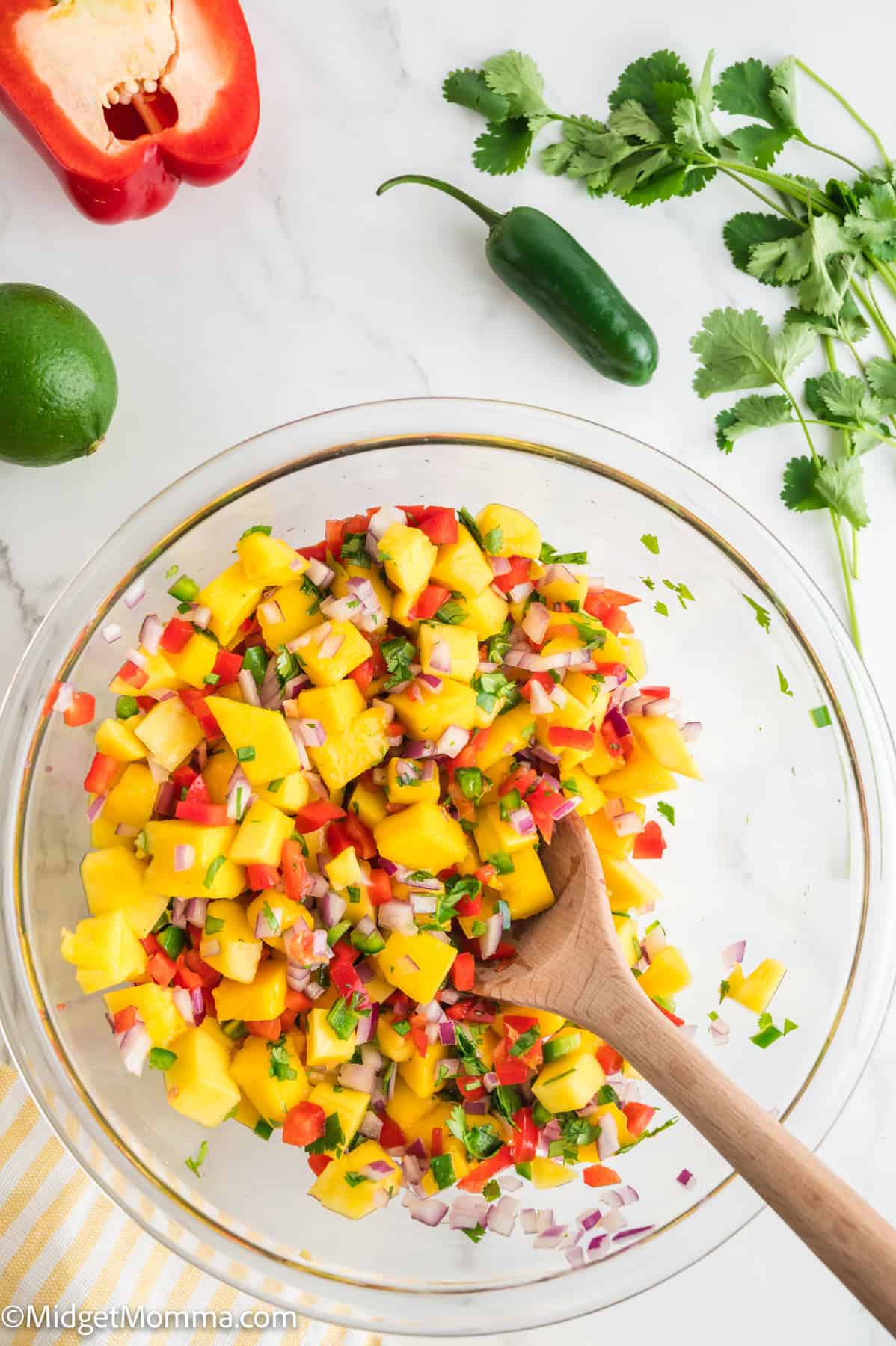 A glass bowl of mango salsa with chopped red onion, red pepper, cilantro, and jalapeno, mixed with a wooden spoon. Fresh ingredients are placed around the bowl.