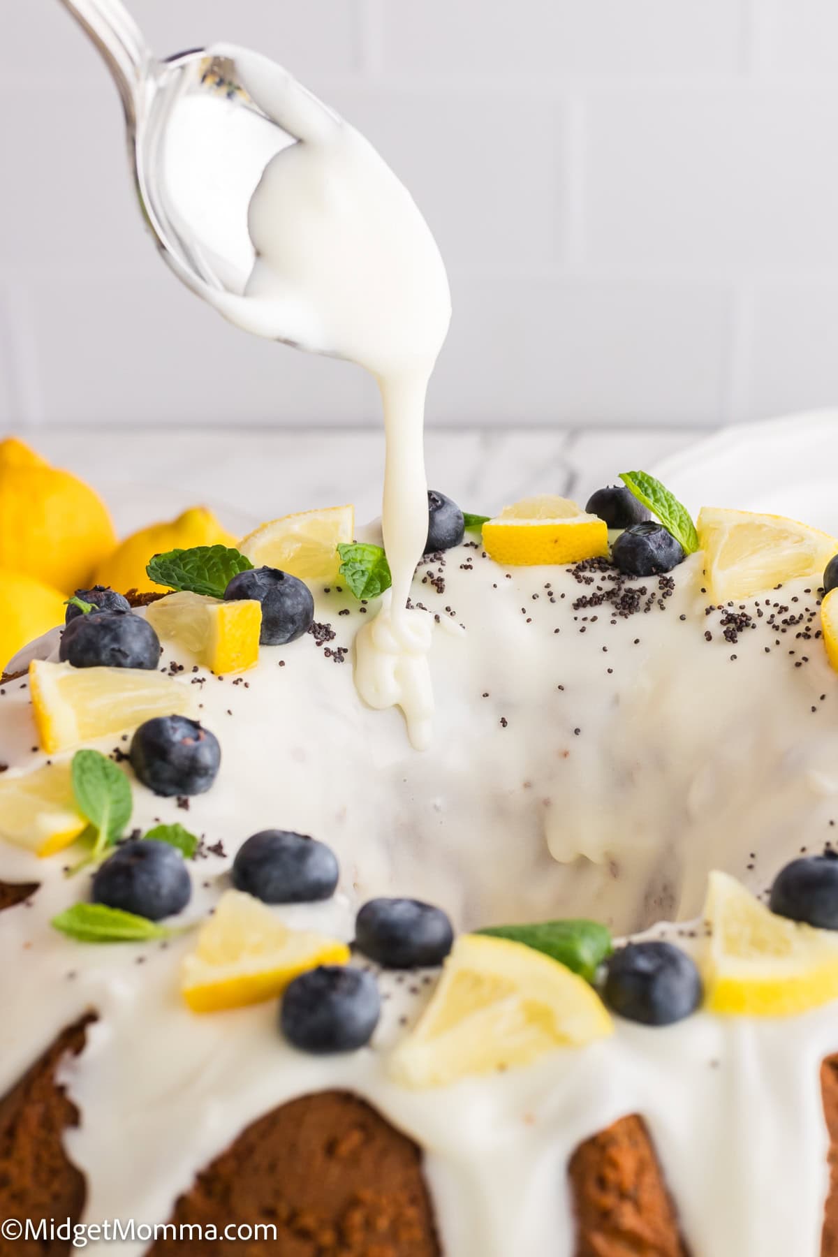 A spoon drizzles icing over a bundt cake topped with blueberries, lemon slices, mint leaves, and poppy seeds.