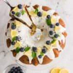 A lemon blueberry bundt cake with white icing, garnished with lemon slices, blueberries, and mint leaves, on a glass stand. Nearby are lemons, a bowl of blueberries, and lemon slices.