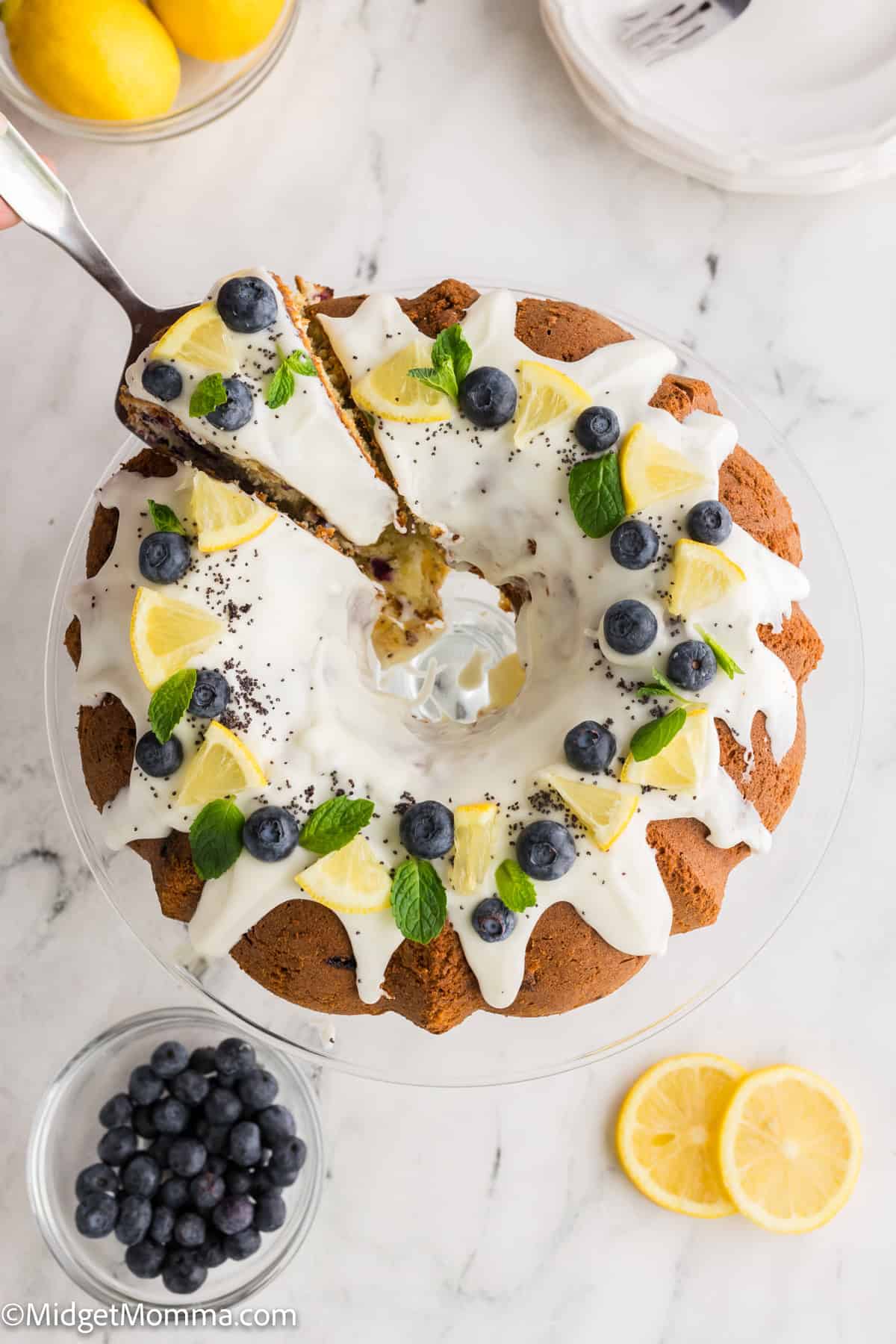A lemon blueberry bundt cake with white icing, garnished with lemon slices, blueberries, and mint leaves, on a glass stand. Nearby are lemons, a bowl of blueberries, and lemon slices.