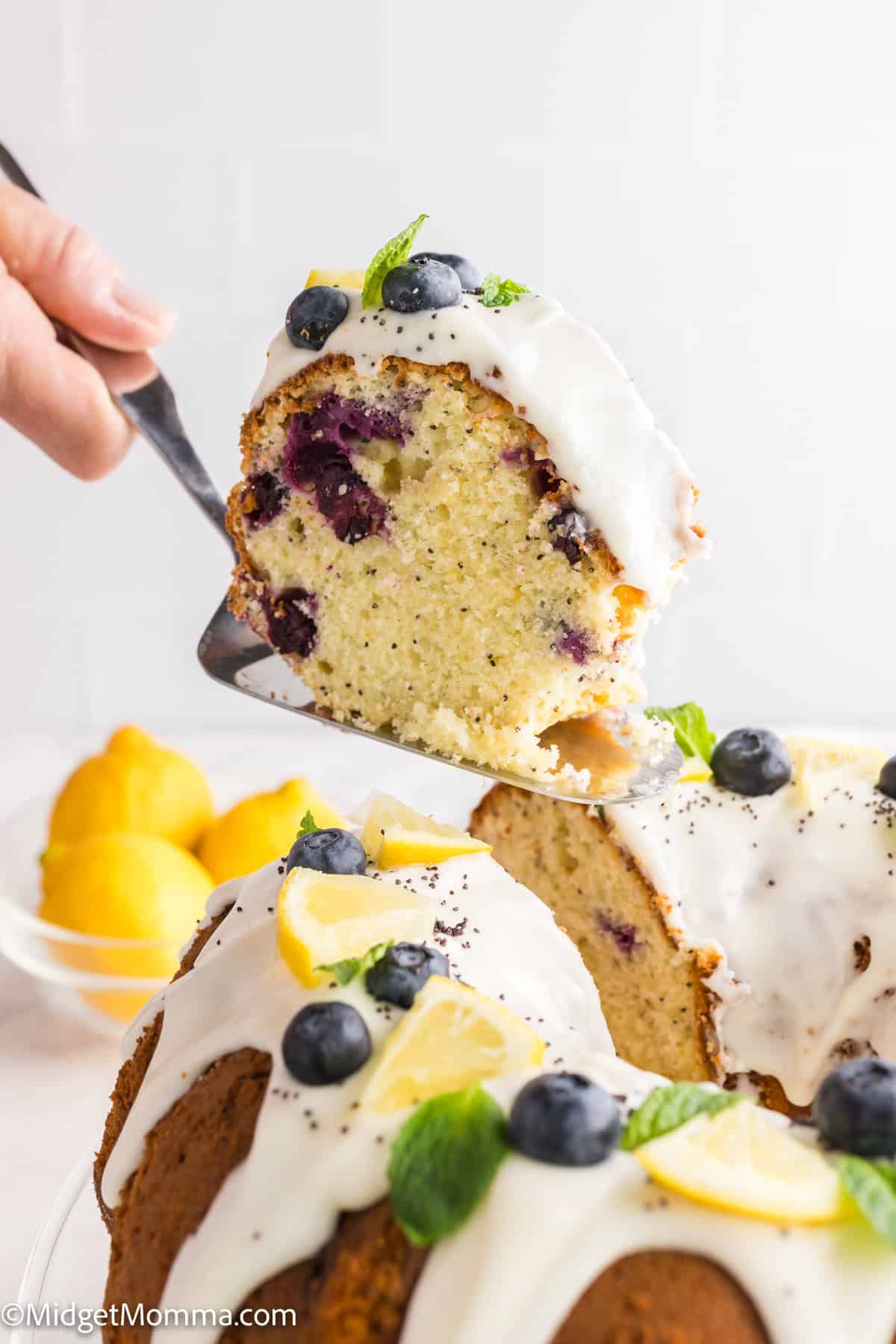 A slice of lemon blueberry bundt cake with white icing, garnished with blueberries, lemon slices, and mint, being lifted from the whole cake.