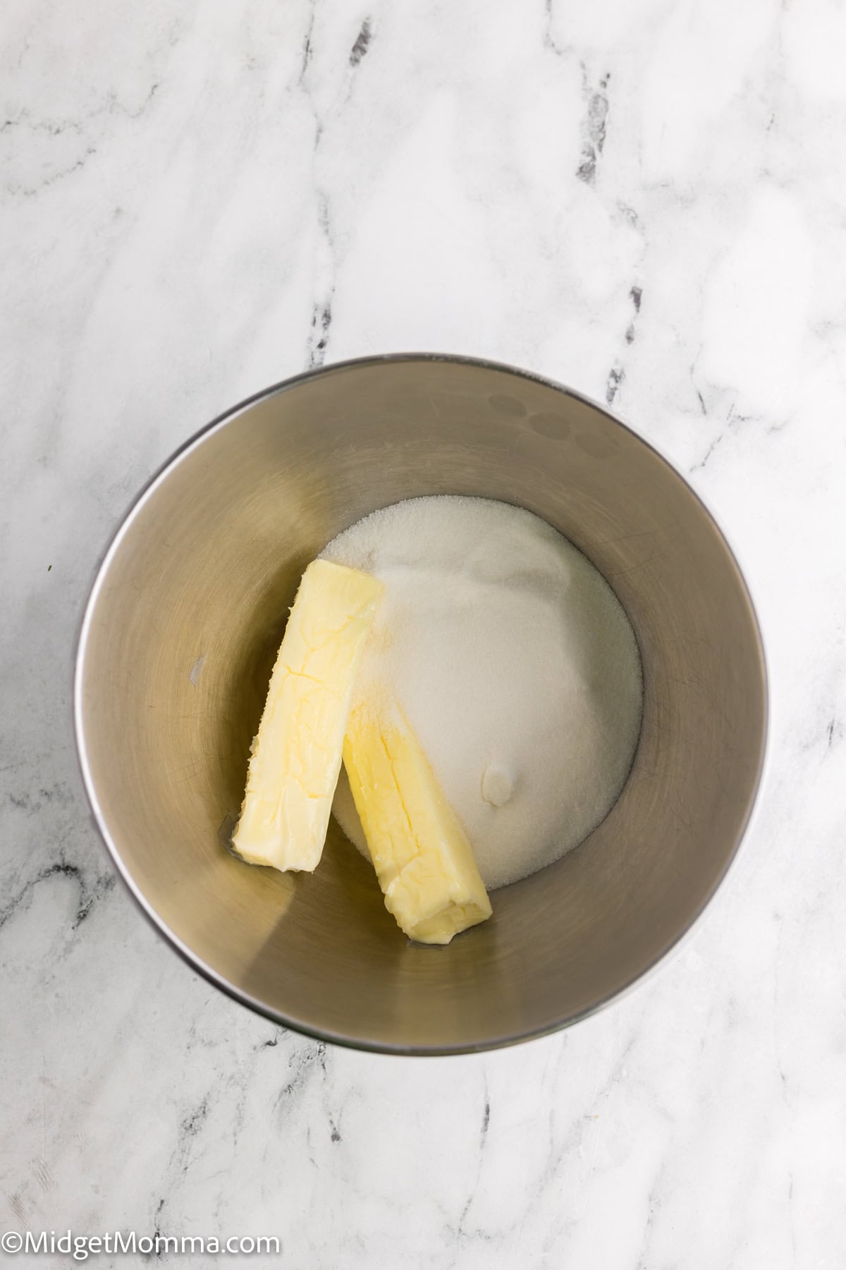 Two sticks of butter and a portion of sugar in a metal mixing bowl on a marble surface.