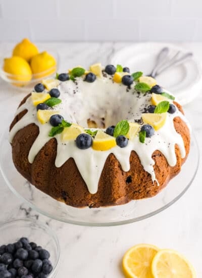 Lemon bundt cake with white glaze topped with blueberries, lemon slices, and mint leaves on a glass stand.