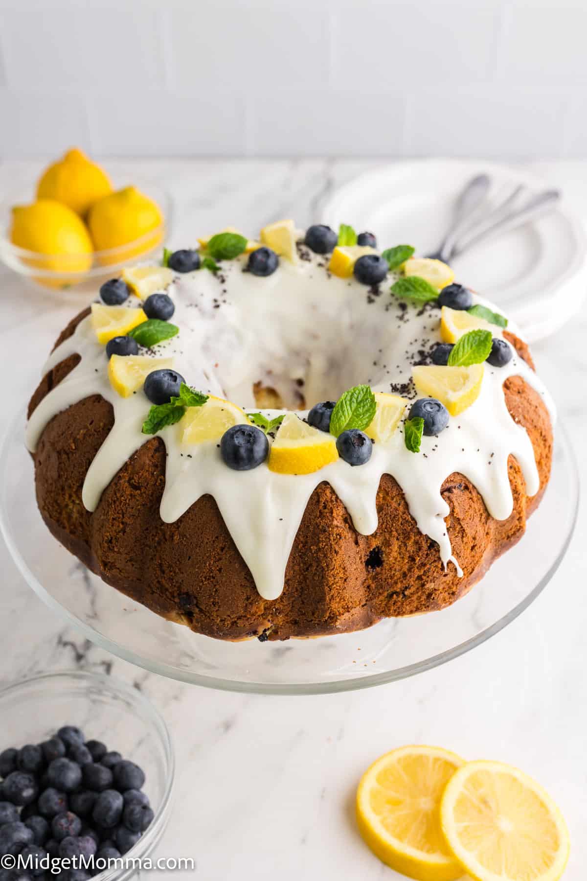 Lemon bundt cake with white glaze topped with blueberries, lemon slices, and mint leaves on a glass stand.