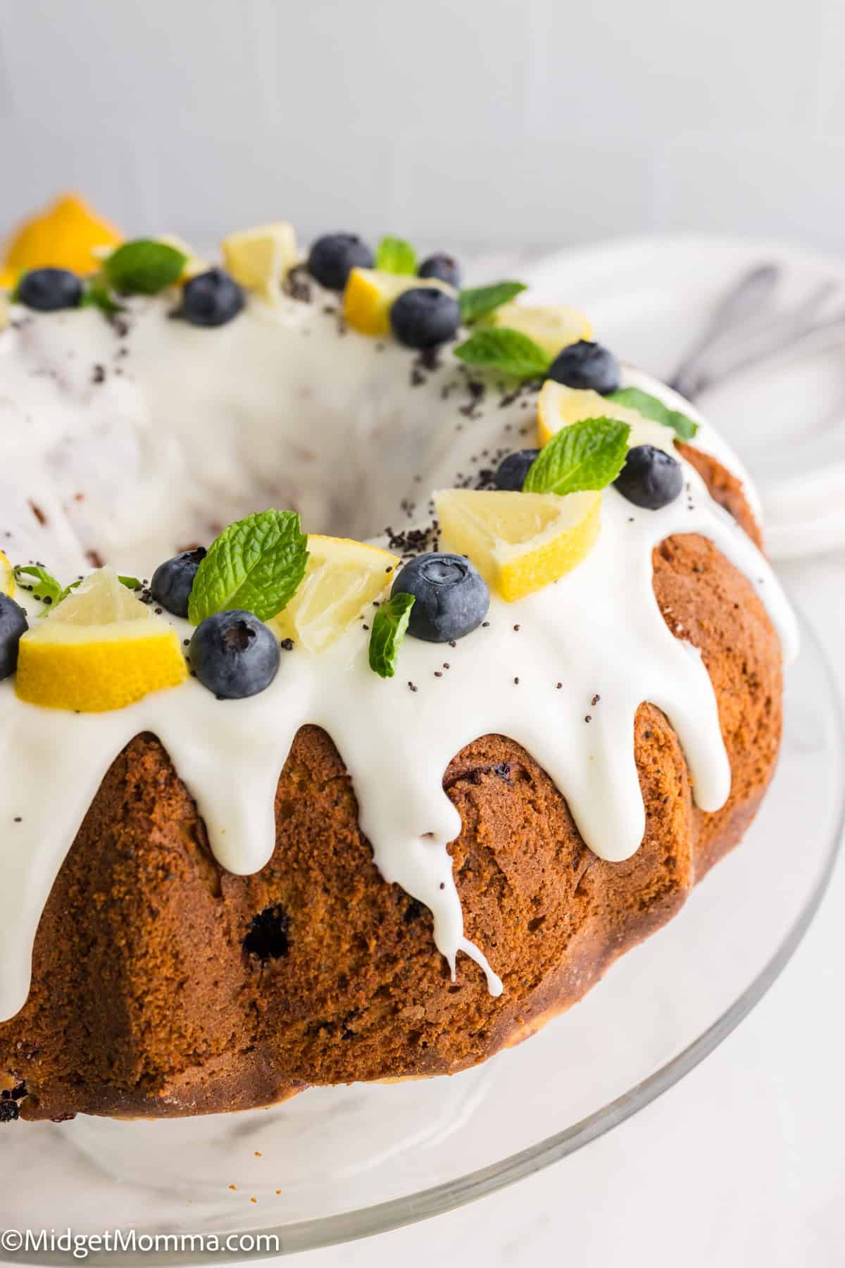 Bundt cake with white icing, topped with blueberries, lemon slices, and mint leaves on a glass stand.